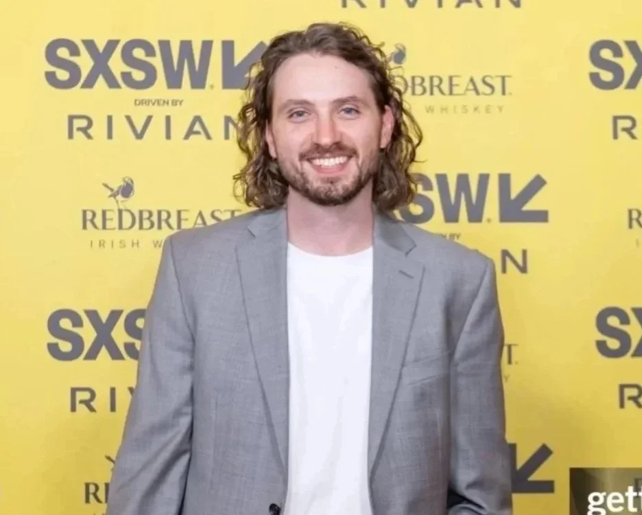 A man with shoulder-length curly hair, a beard, and mustache, wearing a gray blazer and white shirt, standing in front of a yellow backdrop featuring multiple logos for SXSW and other brands.