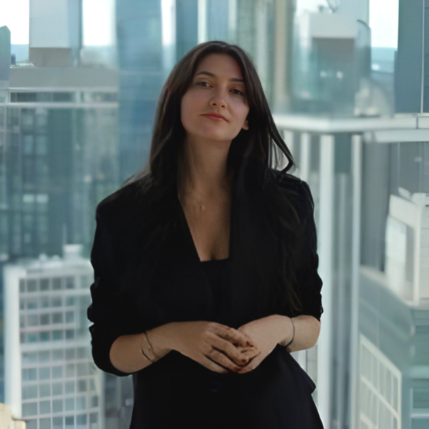 A woman with long dark hair dressed in black, standing in front of large windows in a modern building, looking at the camera with a slight smile.