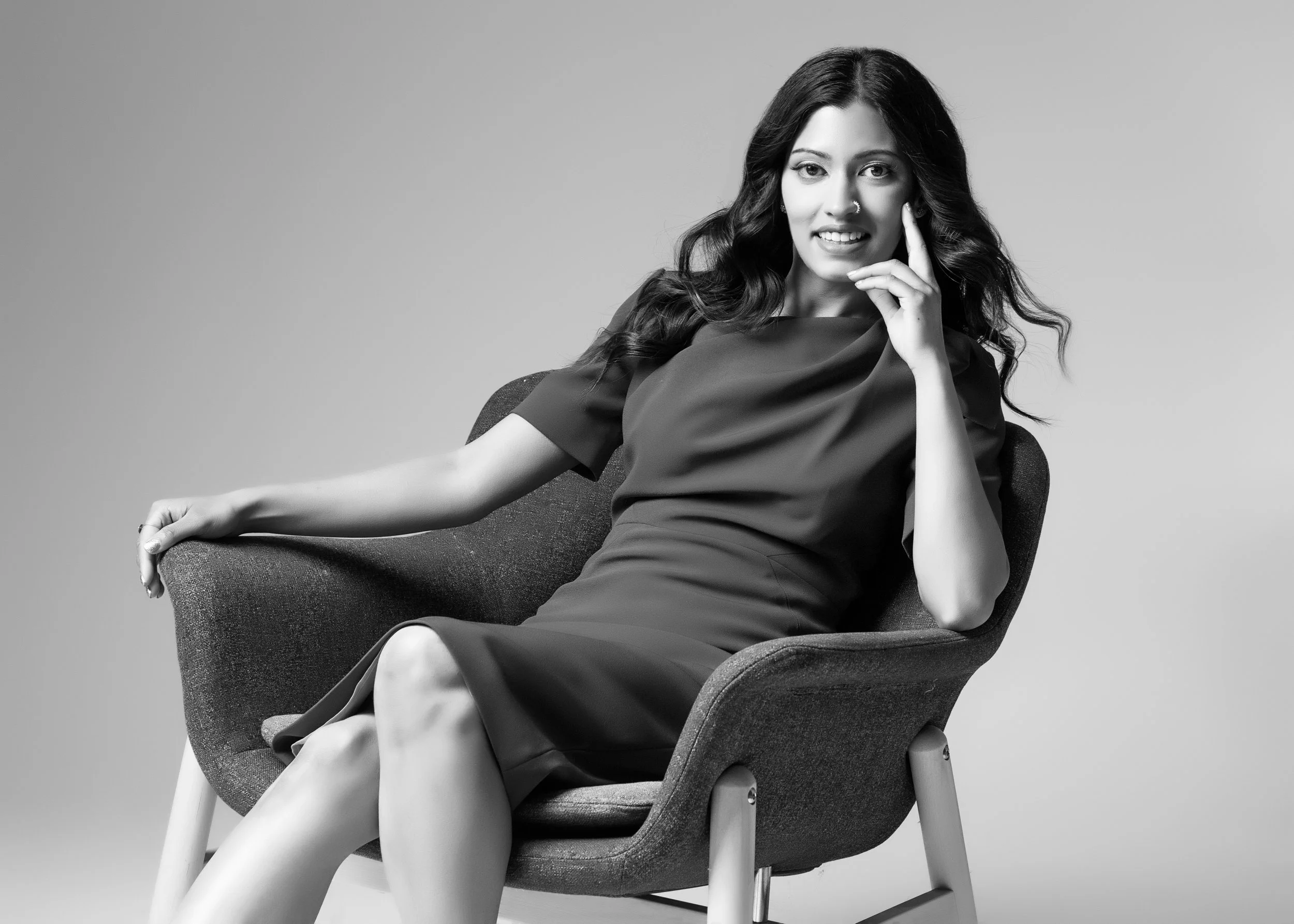 Black and white photo of a woman with dark wavy hair, sitting on a modern chair, smiling, with one hand near her face, wearing a short-sleeved dress.