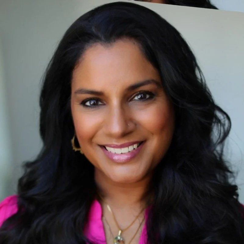 Close-up portrait of a woman with long, dark hair, smiling, wearing gold hoop earrings, a nose ring, a layered necklace, and a pink top.