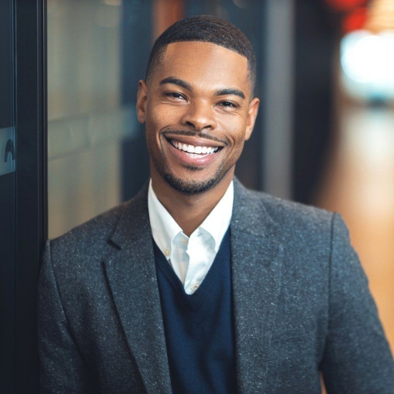 A smiling man in a business suit leaning against a wall in a modern office corridor.
