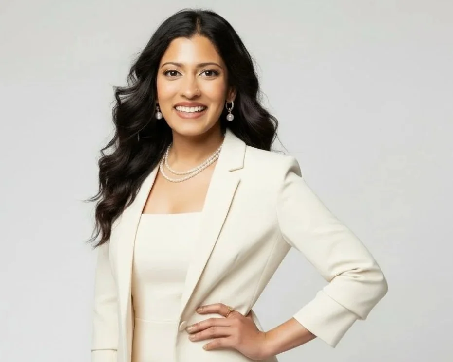 A woman with long dark hair, wearing a white blazer and matching top, accessorized with pearl earrings and a pearl necklace, smiling with a hand on her hip against a plain light grey background.
