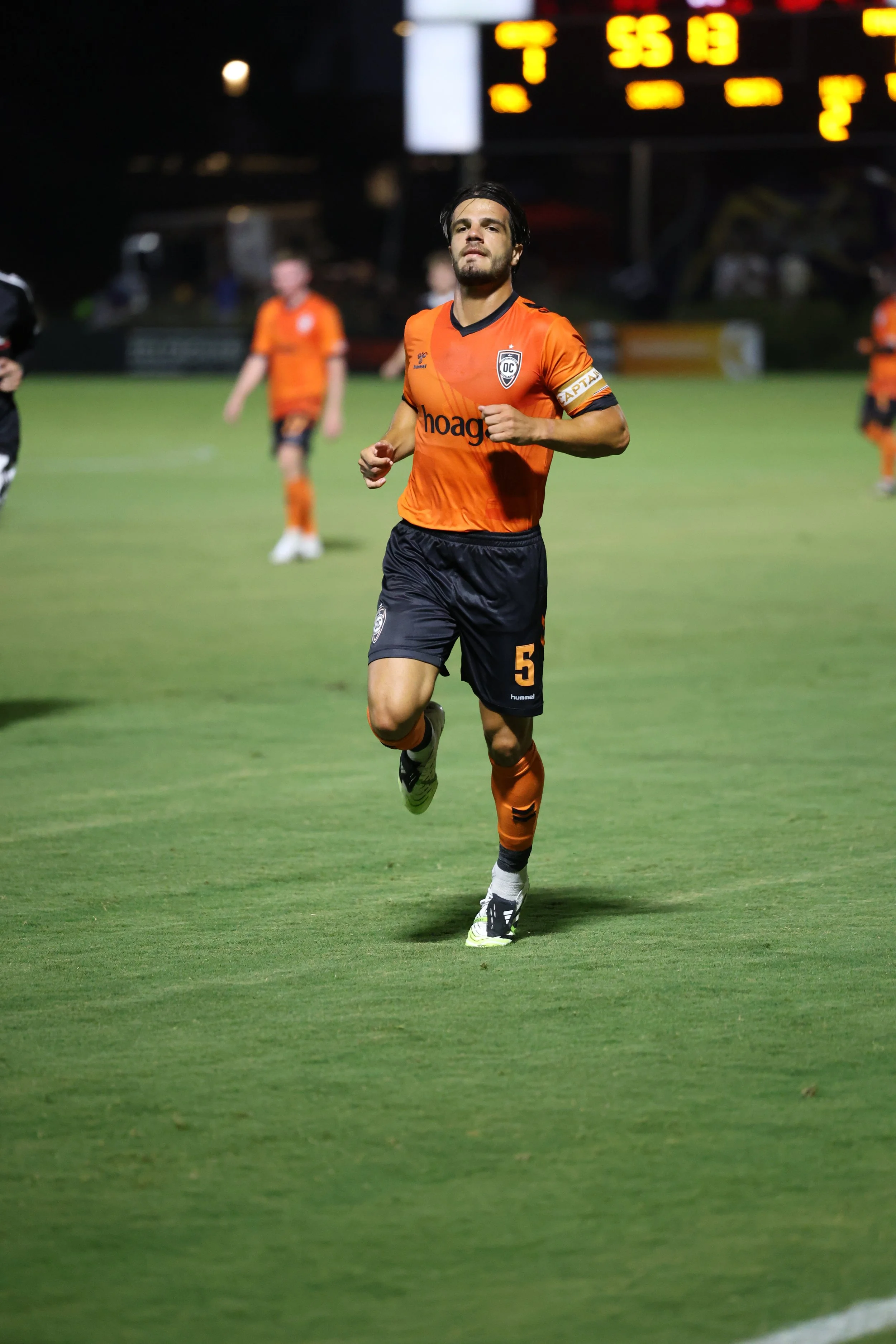 A soccer player in an orange and black uniform running on the field during a night game.