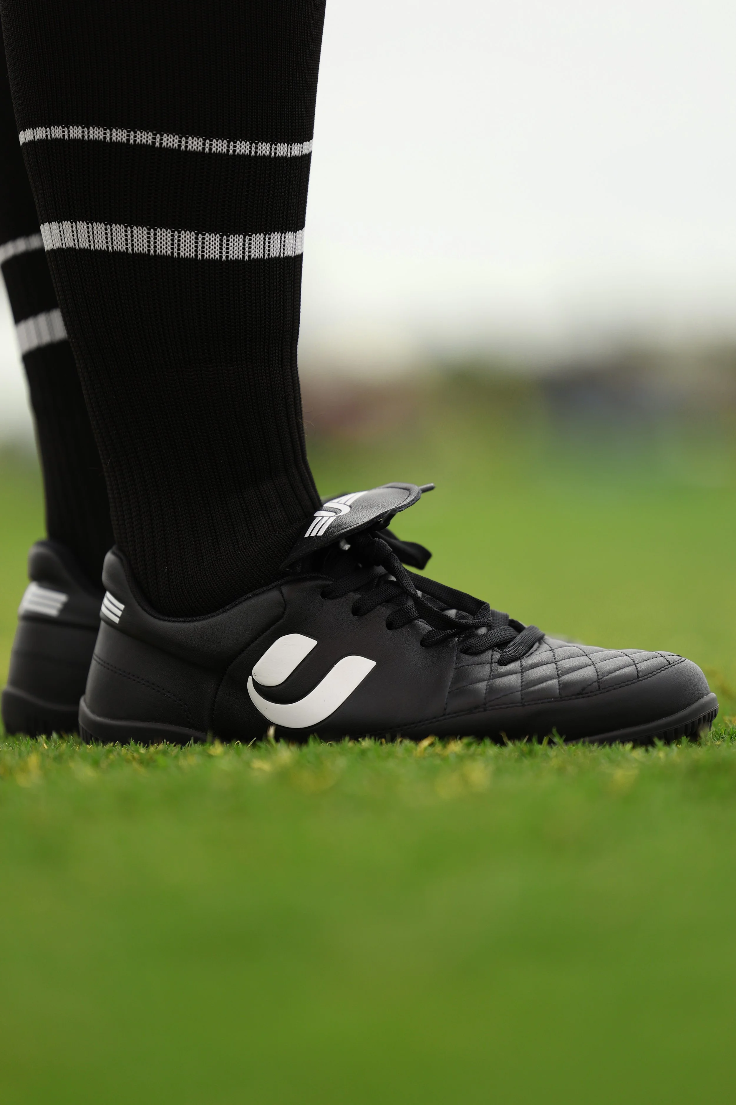 Close-up of a soccer player's black cleats and black socks with white stripes, standing on green grass.