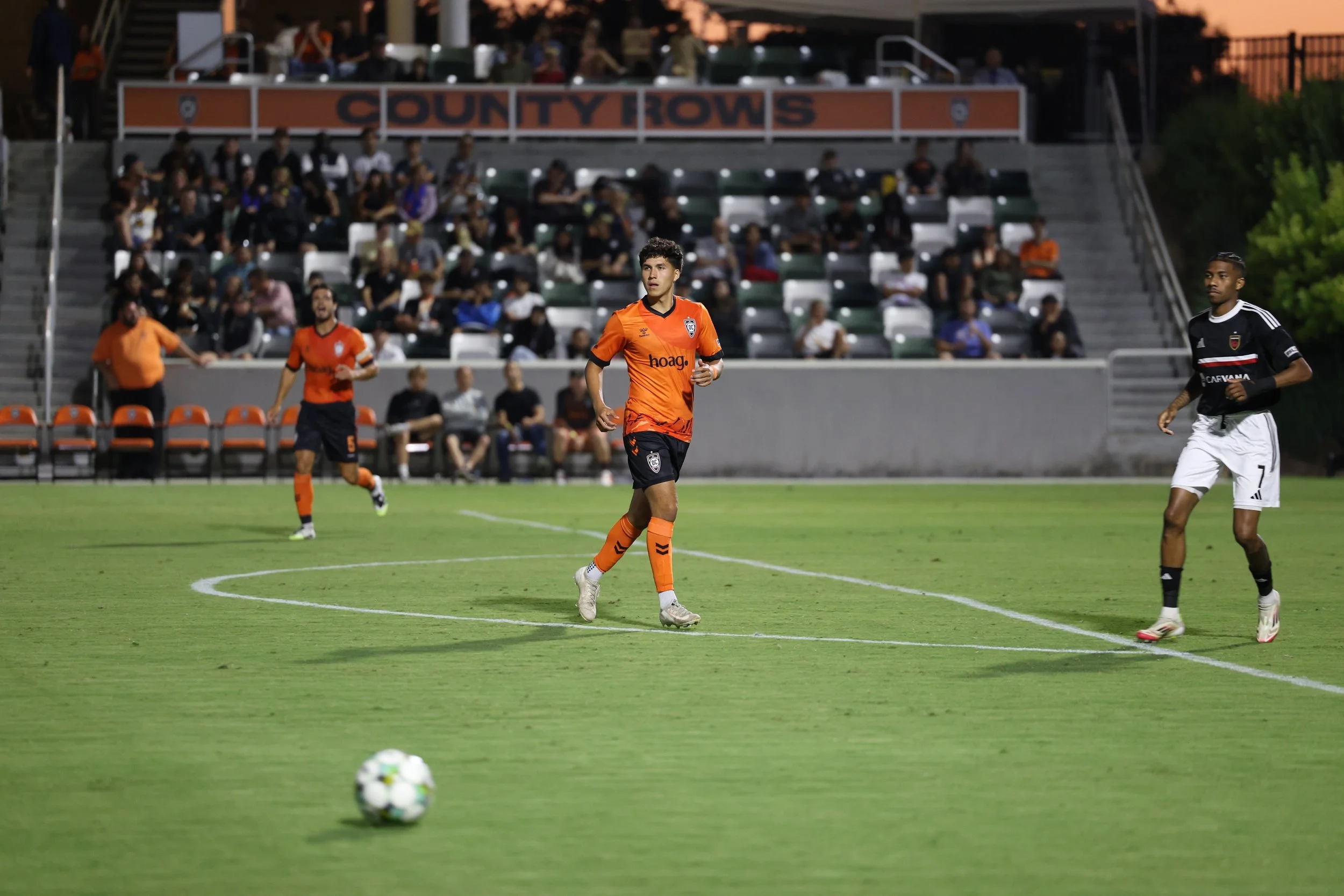 Soccer players on the field during a game, with spectators watching from the stands in the background.