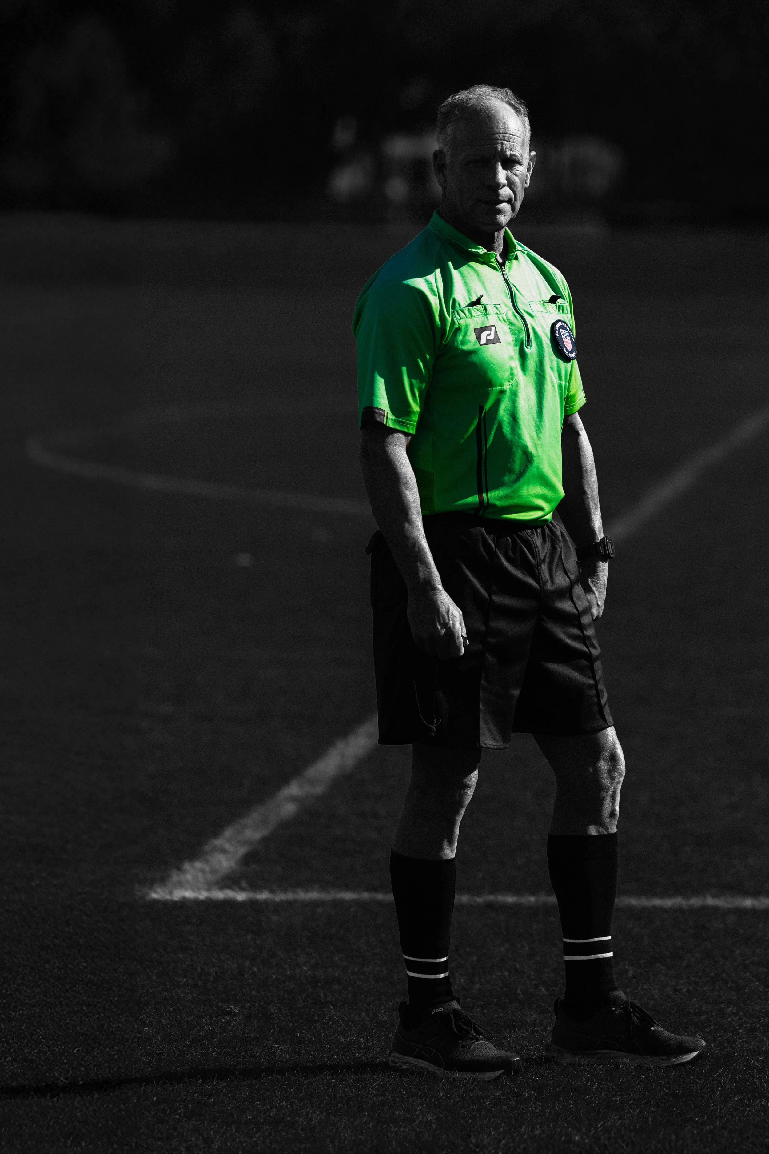 A grayscale image of a soccer referee wearing a chloe green shirt and black shorts, standing on a soccer field.