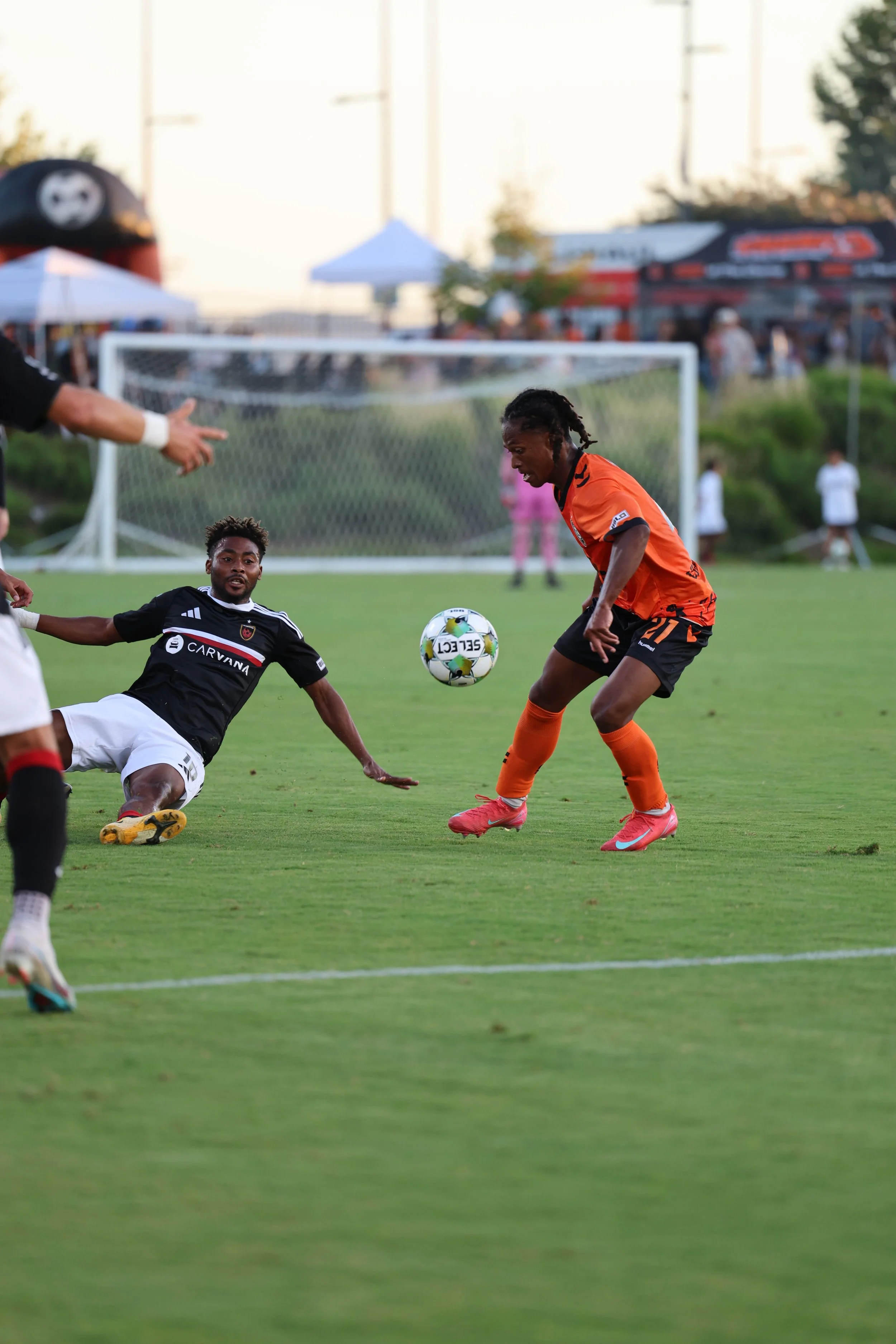 Two soccer players, one in black and one in orange, compete for the ball during a game on a green field, with a goal in the background.