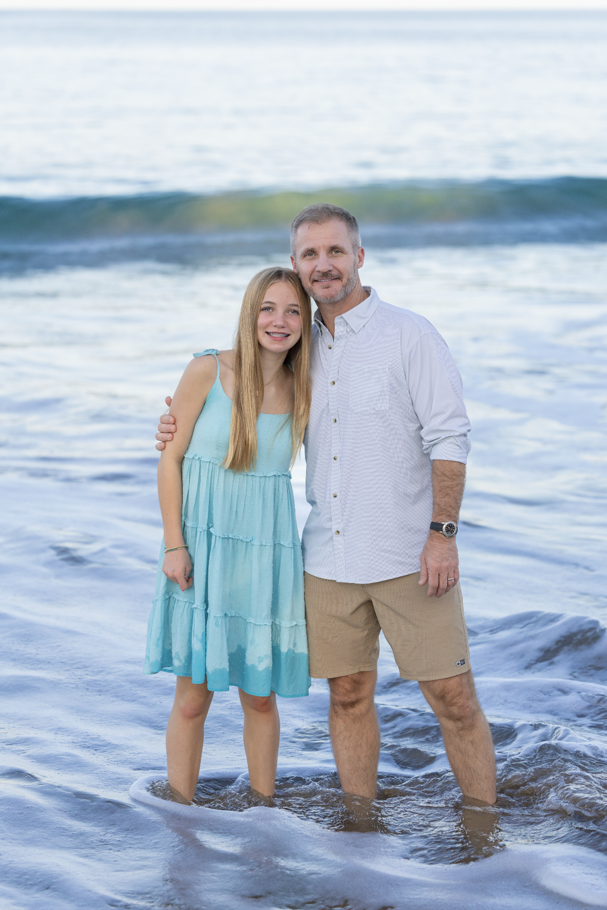 A father and daughter standing in the ocean with waves behind them, smiling for their photographer Cynthia Buckles with CynthiArtistry. Cynthia Buckles is rated best photographer on the Big Island of Hawaii near Waimea, Waikoloa and Kona.