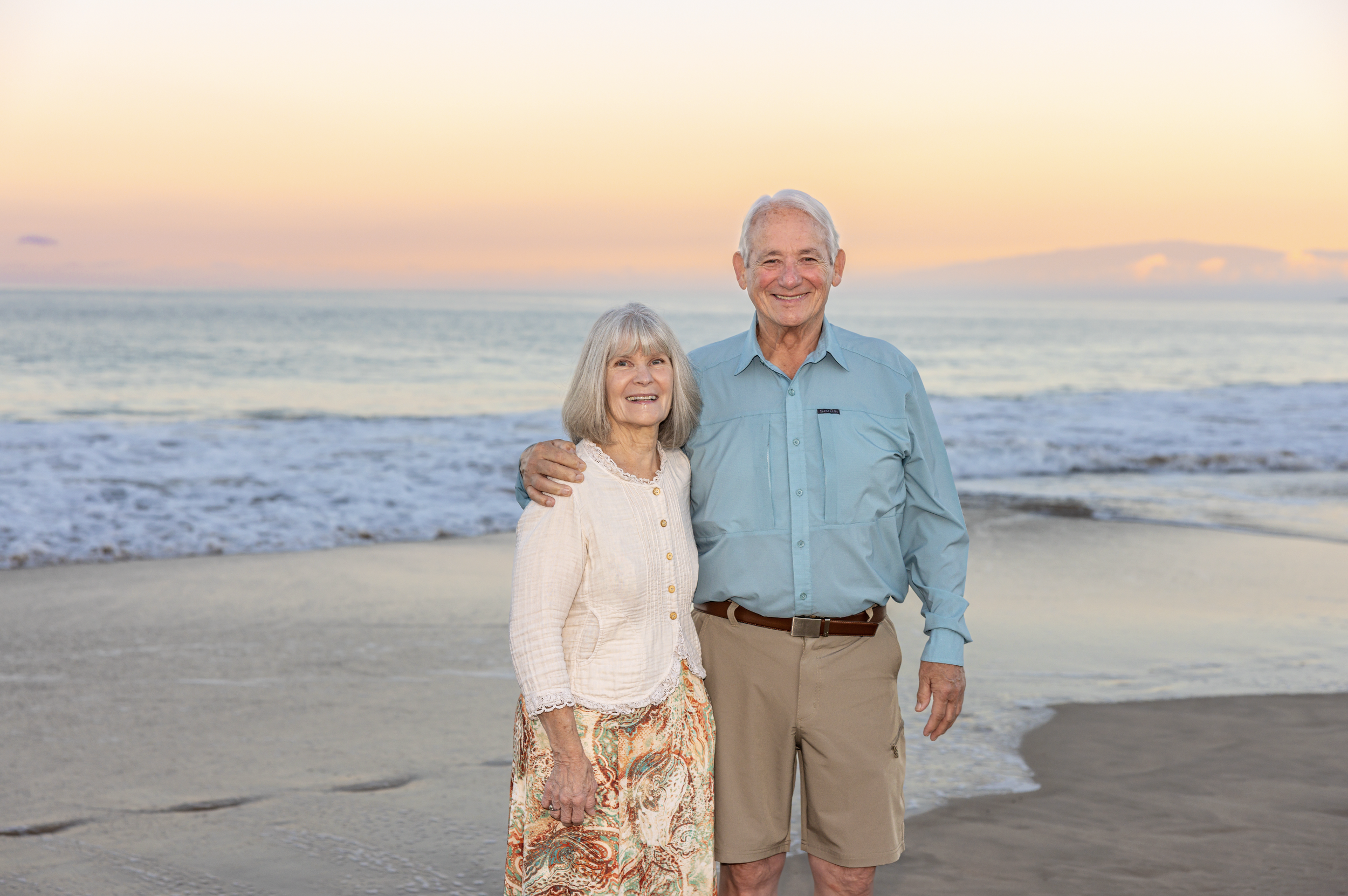 An elderly couple stands on the beach at sunrise laughing with joy at Hapuna Beach on the Big Island of Hawaii near the Mauna Kea Hotel and Westin Hapuna Hotel with their wonderful photographer Cynthia Buckles, the best photographer in Hawaii!