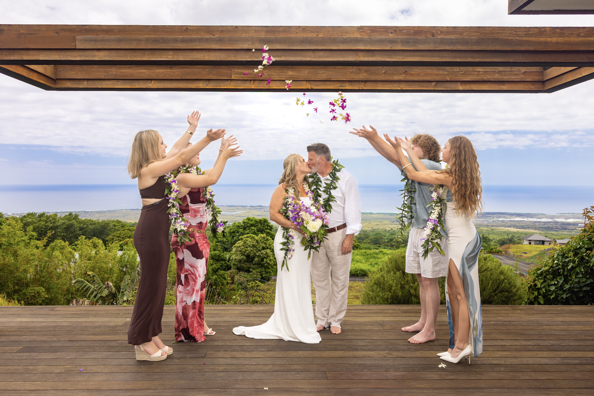 A wedding ceremony with a couple kissing and exchanging vows on a wooden deck outdoors, surrounded by friends throwing flower petals, with a scenic landscape of greenery and the ocean in the background. The location is Kailua Kona, Big Island Hawaii.