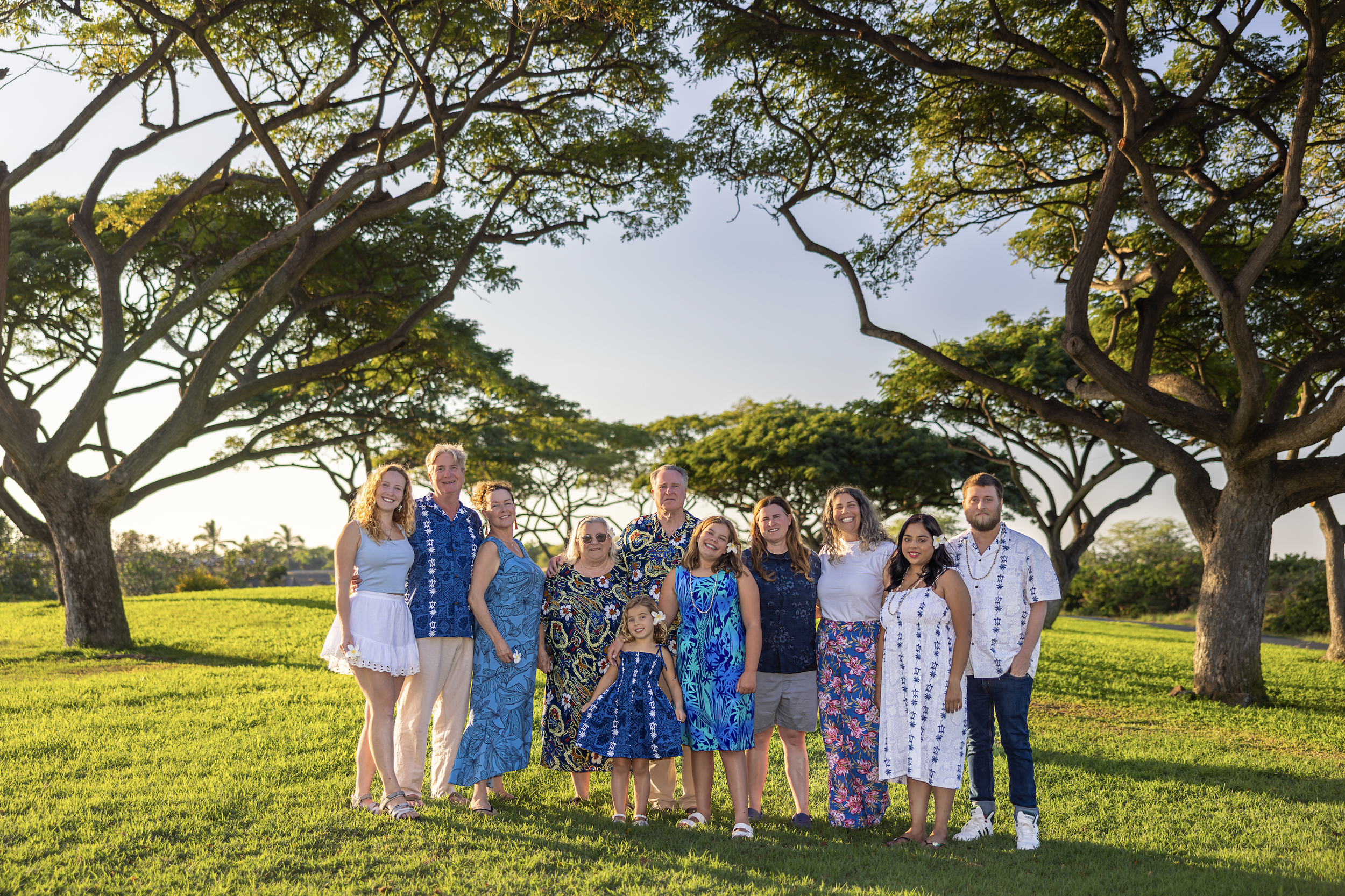 A group of family members standing outdoors on a lush green lawn under large trees in bright sunlight. On the Big Island of Hawaii near Kailua Kona with their wonderful photographer Cynthia Buckles, rated the best photographer in Hawaii.