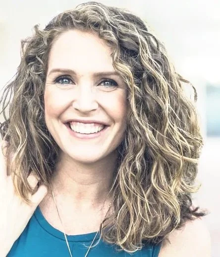 A woman with curly blond hair, blue eyes, and a bright smile wearing a blue sleeveless top.
