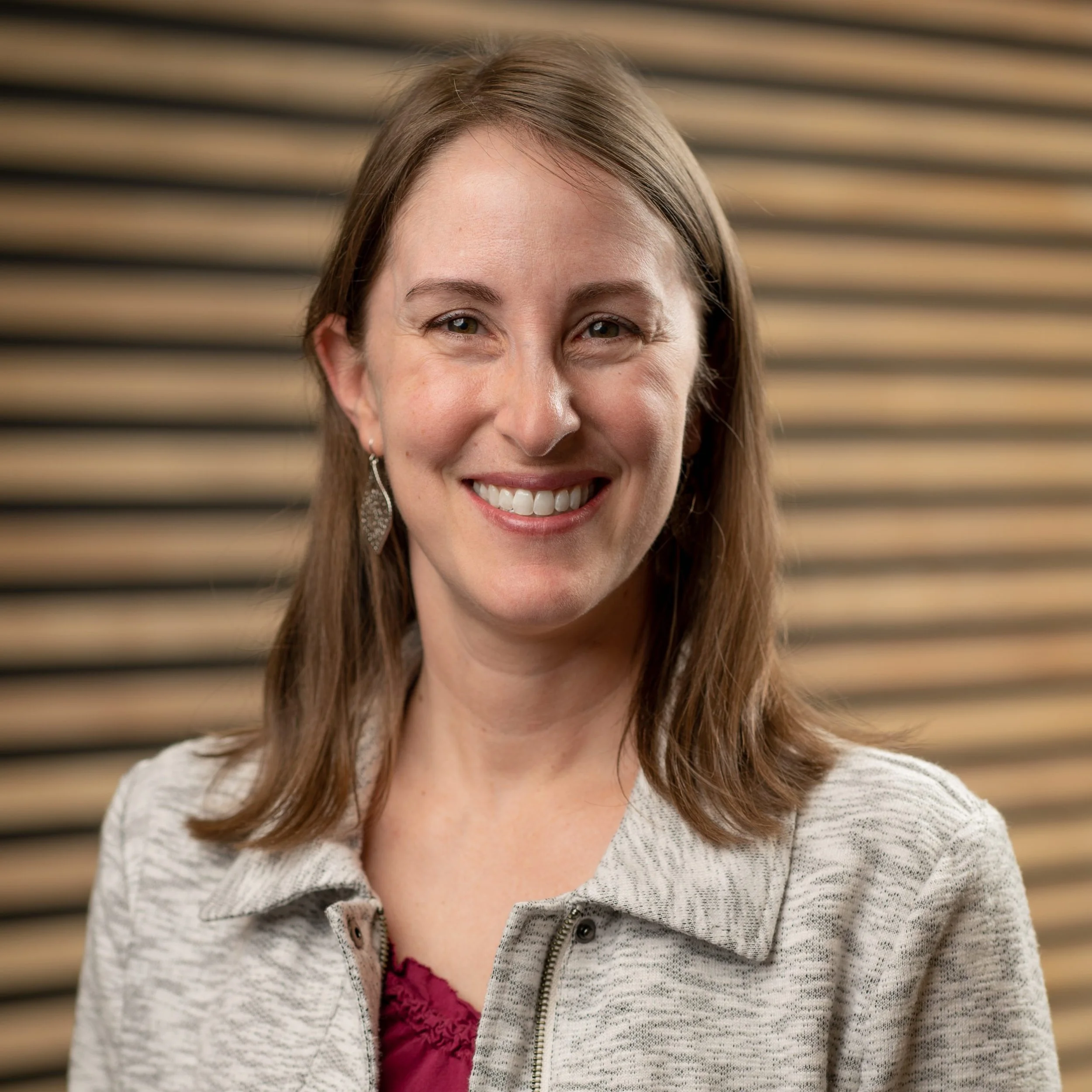 A woman with shoulder-length brown hair, wearing earrings, a beige jacket, and a burgundy top, smiling at the camera against a background of horizontal wooden panels.