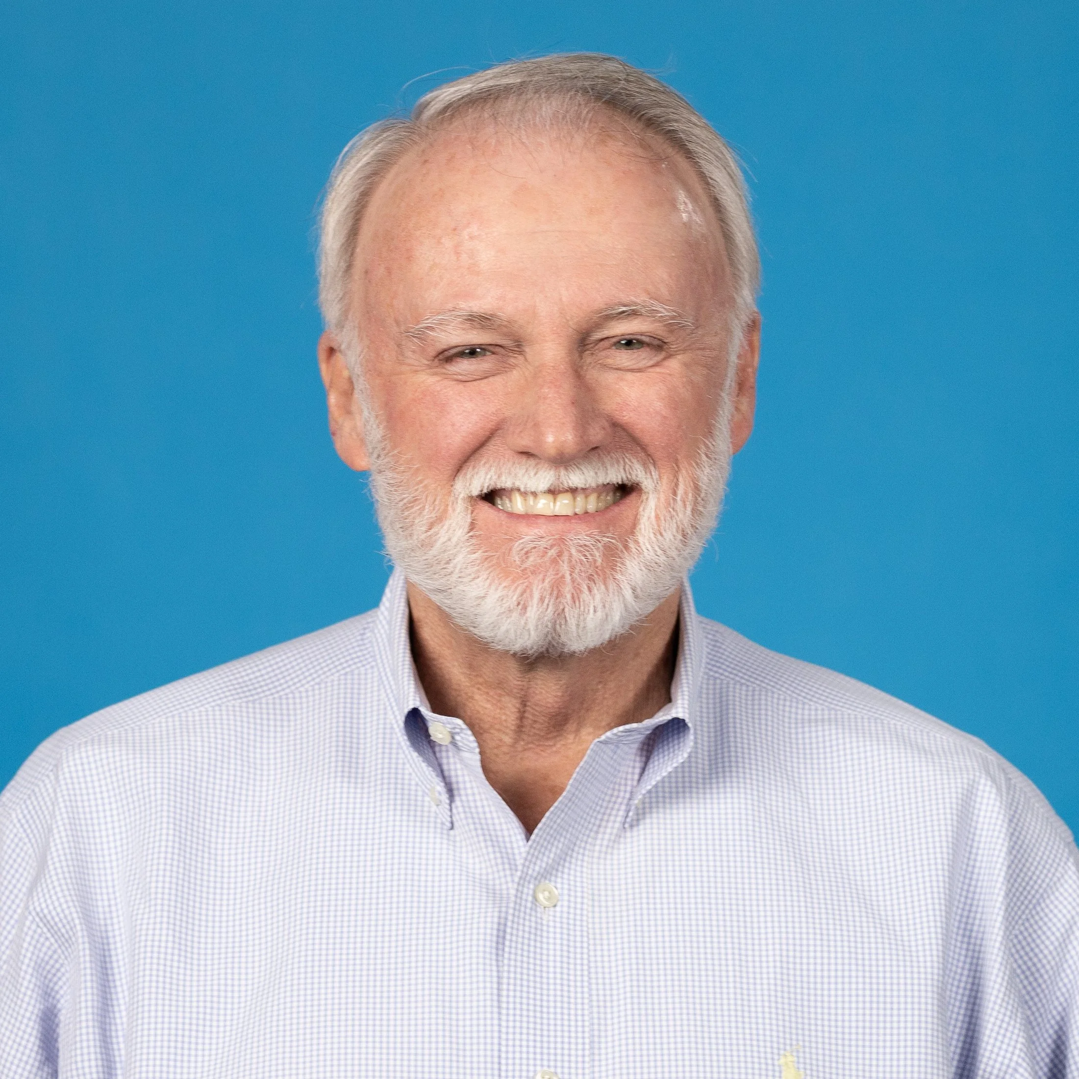 Smiling elderly man with a white beard and gray hair, wearing a light blue checkered dress shirt, against a solid blue background.