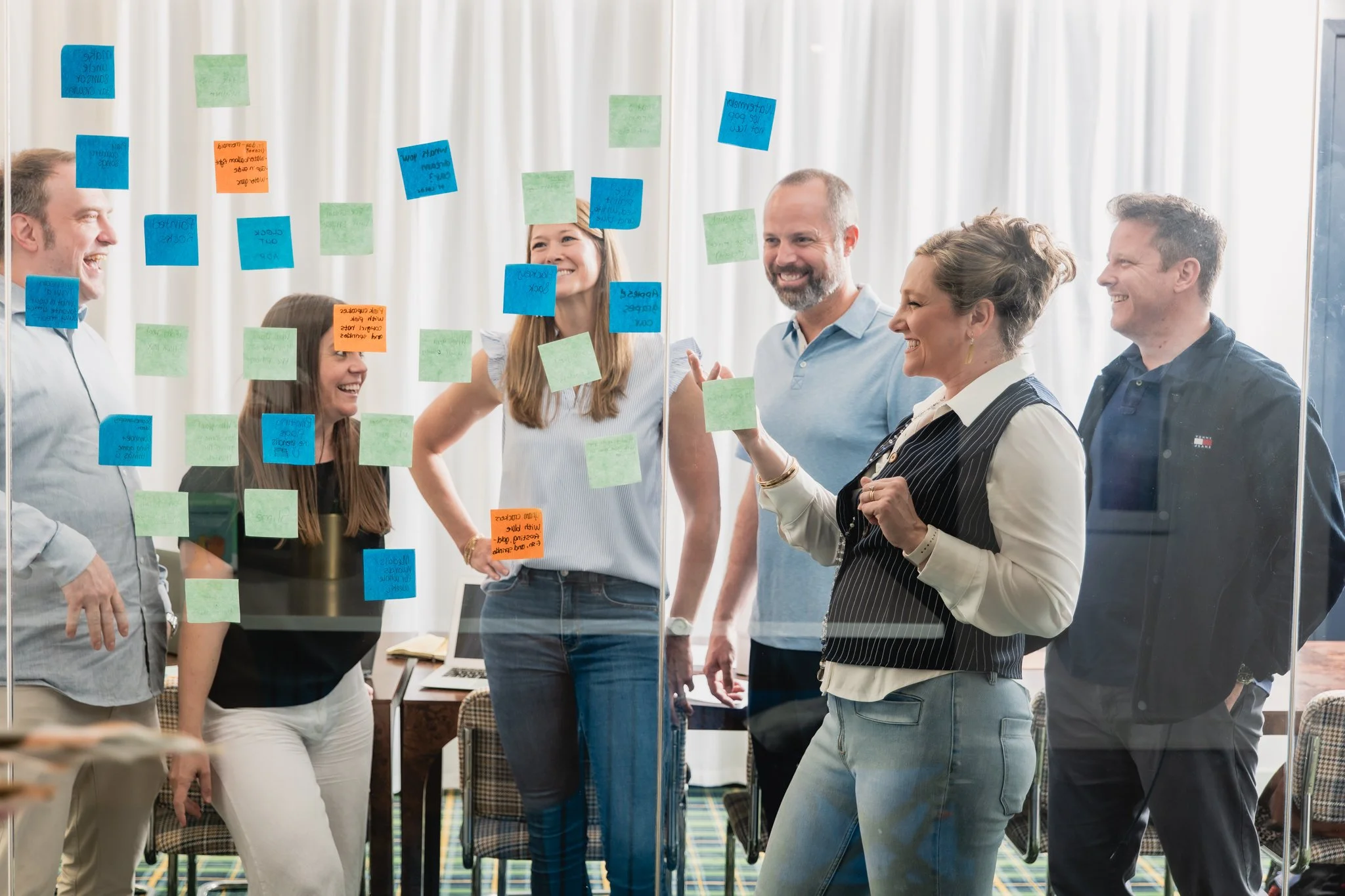 Group of six adults standing around a glass wall covered with colorful sticky notes, smiling and engaging in a brand strategy workshop discussion.