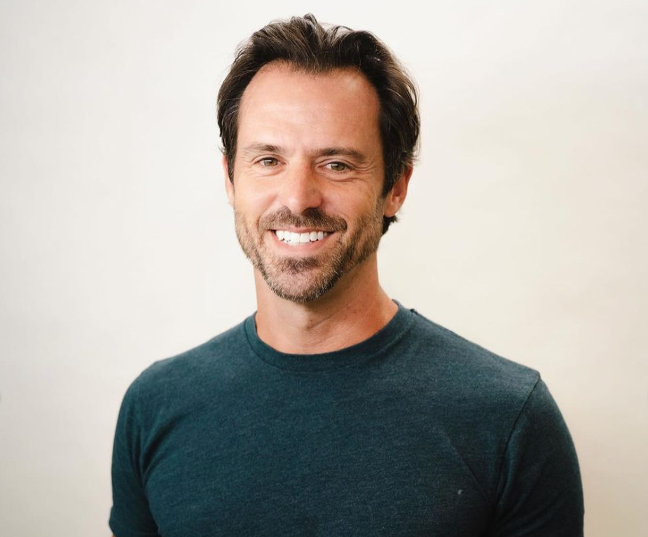 A man with dark hair and a beard, smiling, wearing a dark green t-shirt, standing against a plain white background.