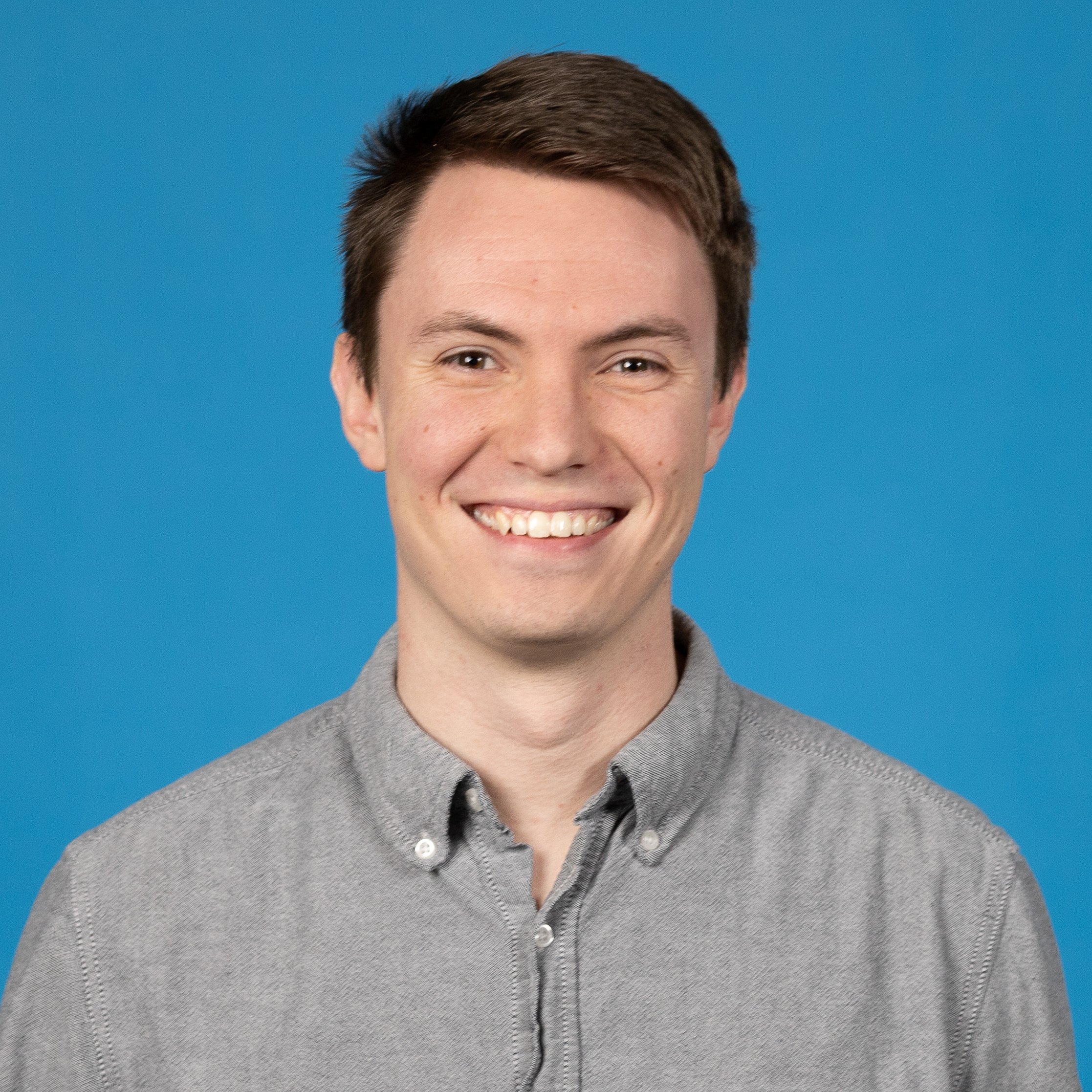 Close-up portrait of a young man with short brown hair, smiling widely, wearing a gray button-up shirt against a blue background.