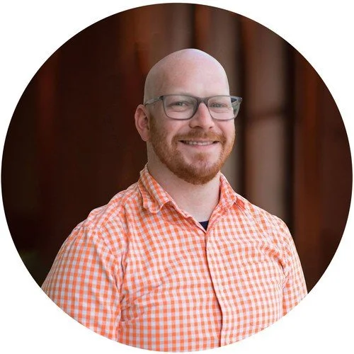 A smiling man with glasses, a beard, and a bald head, wearing an orange checkered shirt, standing in front of a wooden background.