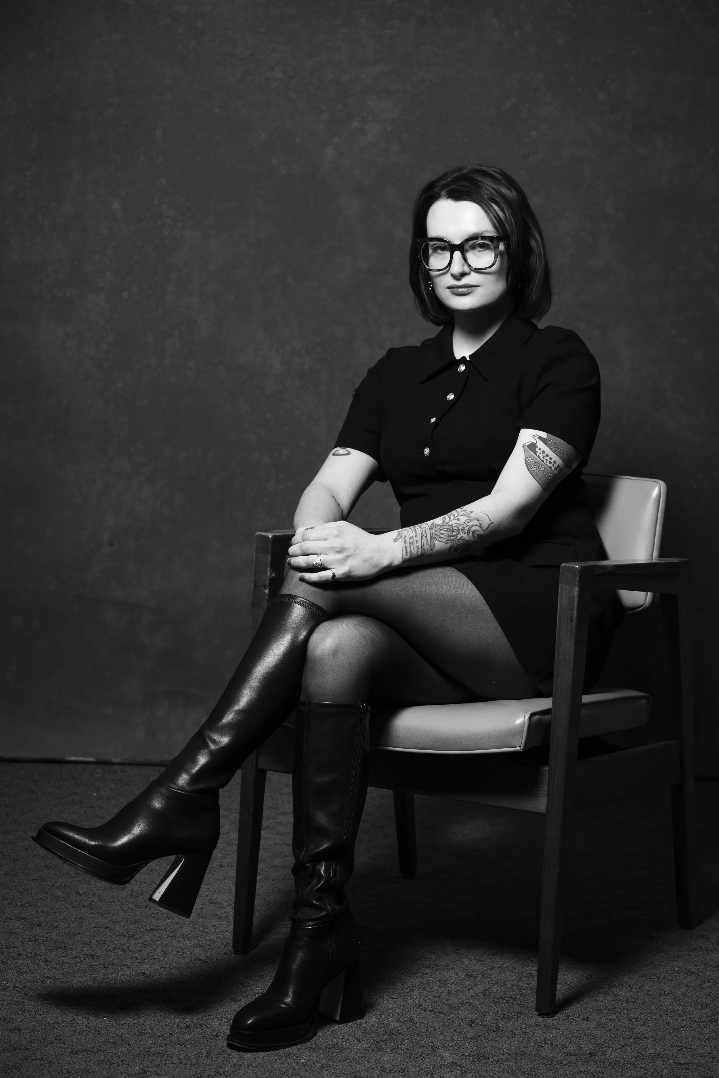 Black and white photo of a woman with glasses sitting cross-legged in a chair, wearing a black dress and high-heeled boots, with tattoos on her arms, against a plain dark background.