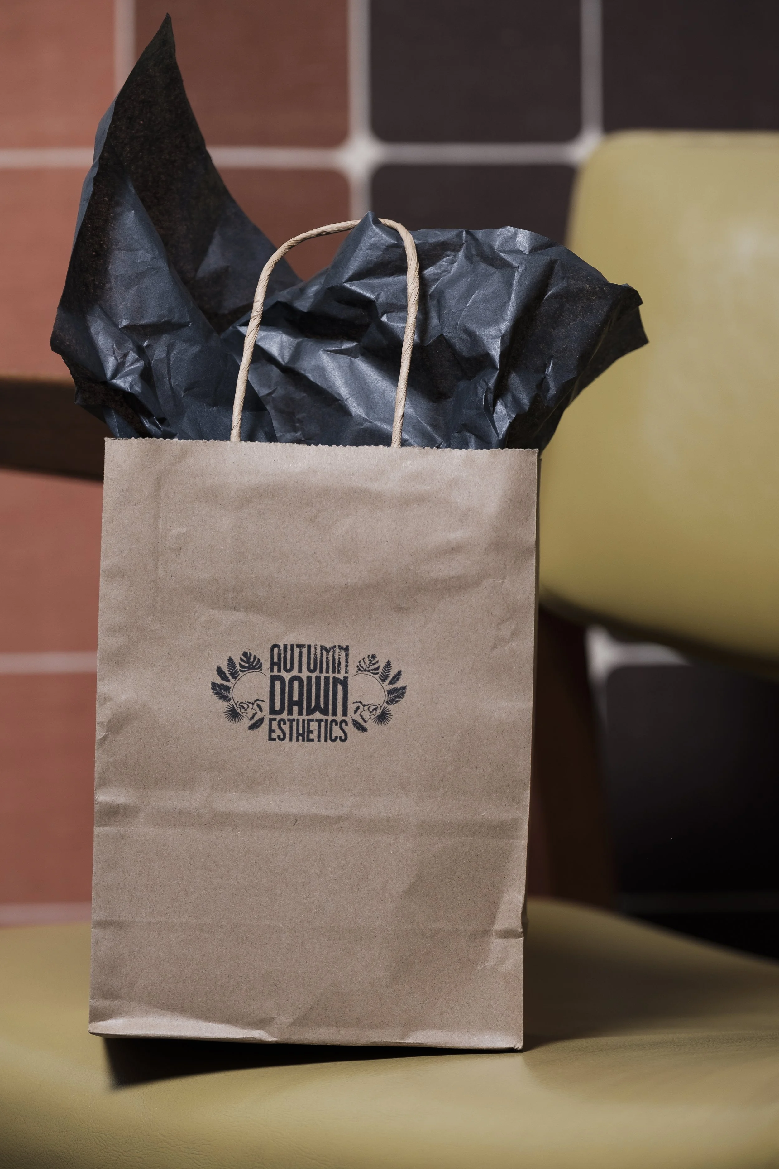 A paper shopping bag labeled 'Autumn Dawn Esthetics' with black crumpled tissue paper inside, placed on a yellow surface with tiled wall in the background.