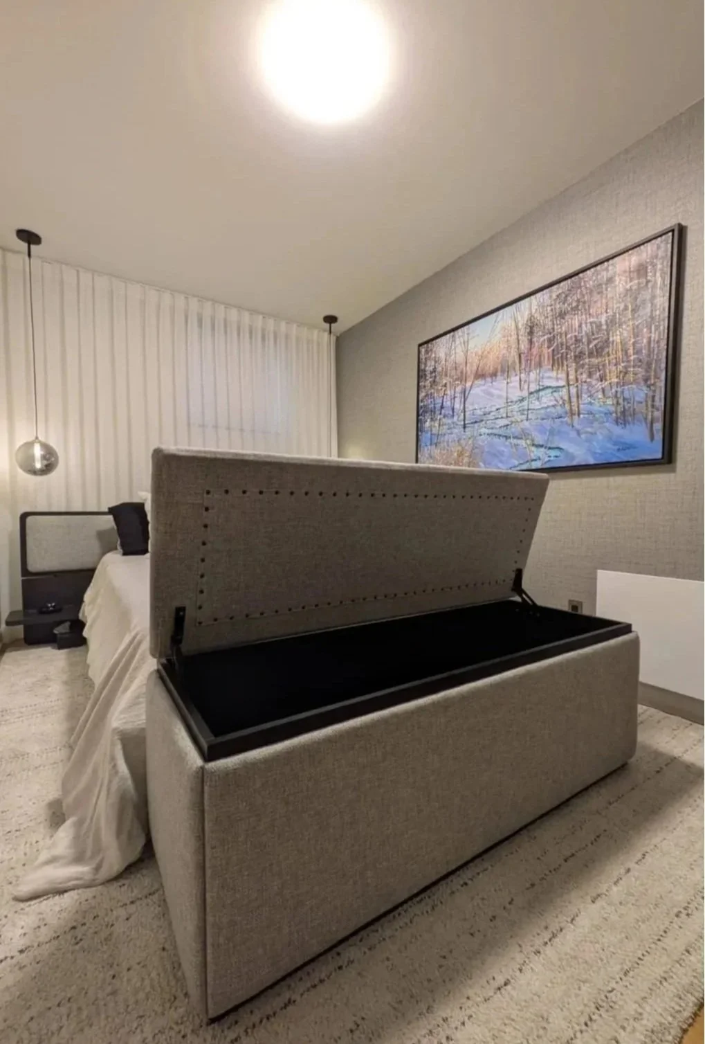 Modern bedroom with an open gray upholstered storage bed, wall-mounted winter landscape artwork, pendant lights, and white curtains