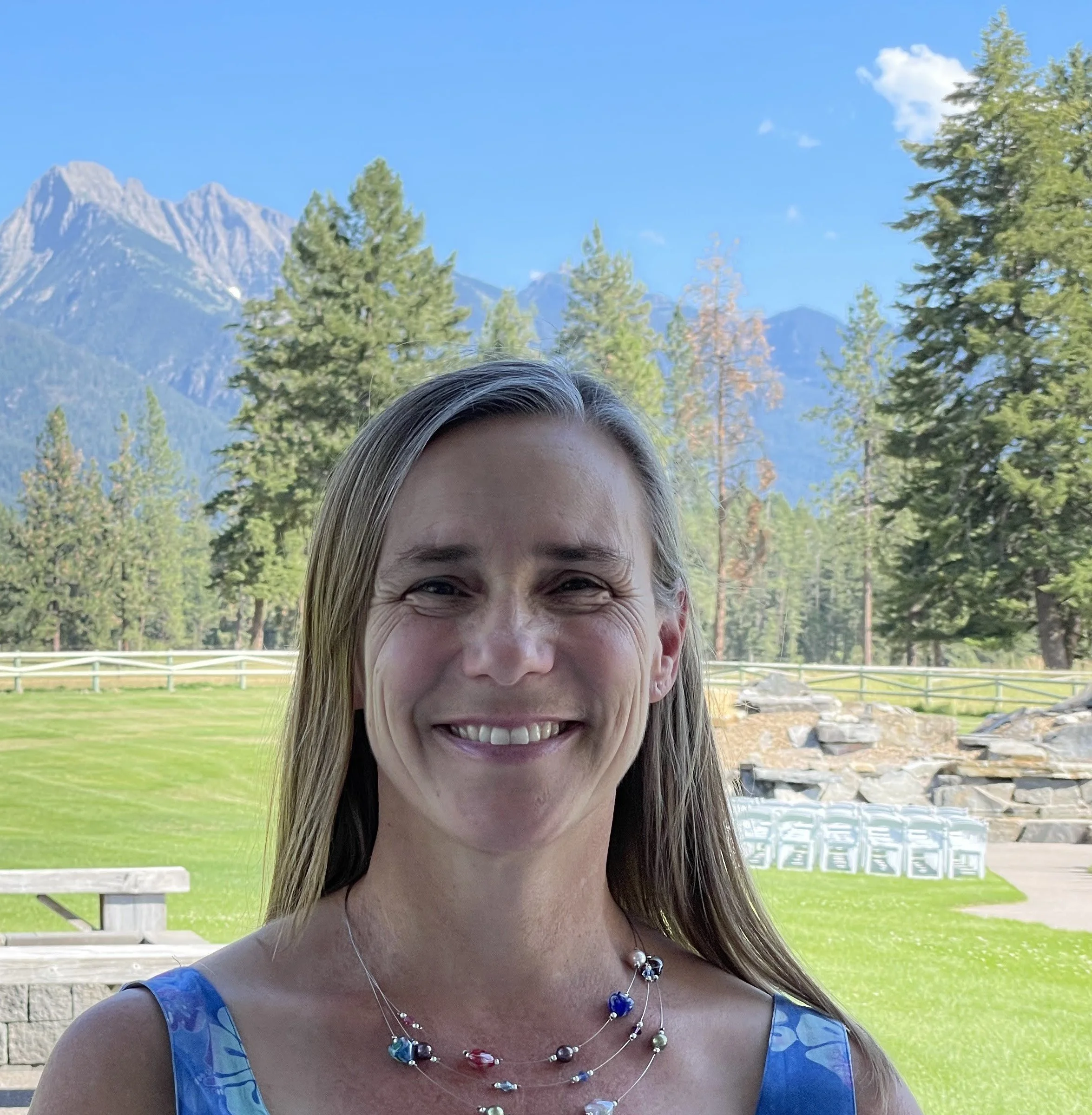 headshot of the therapist, Anne, standing in front of spruce trees and snow-peaked mountains.  The subject is smiling and wearing a blue, floral summer dress.
