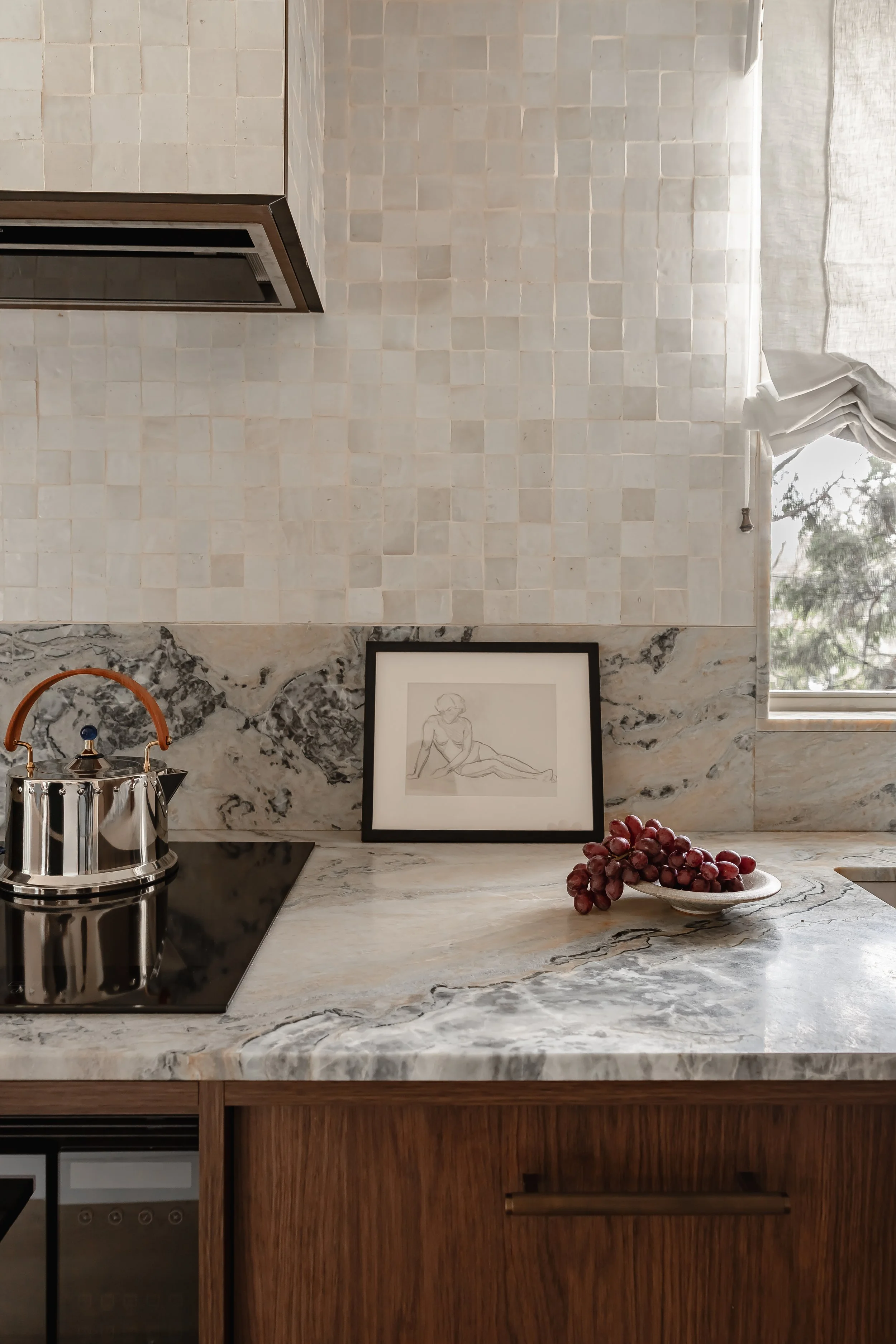 A kitchen with a marble countertop, a framed line art drawing of a woman, a bunch of grapes on a plate, a window with a curtain, a stainless steel kettle, and wooden cabinets.