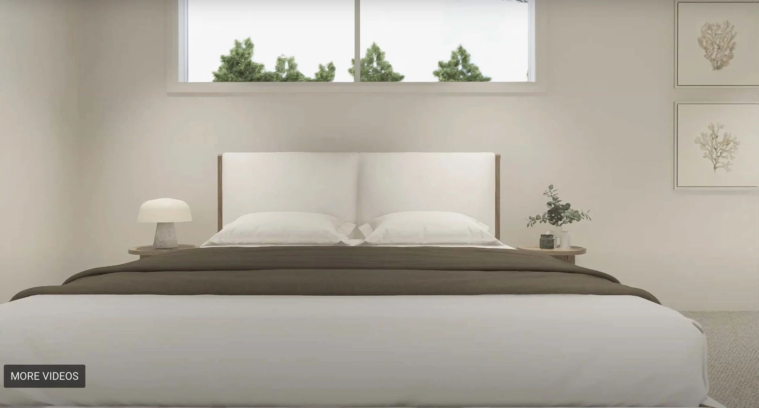 A modern bedroom with a white bed and a wooden headboard, a small table lamp, and some decorative plants on a nightstand. There is a window with trees outside and wall art on the right side.