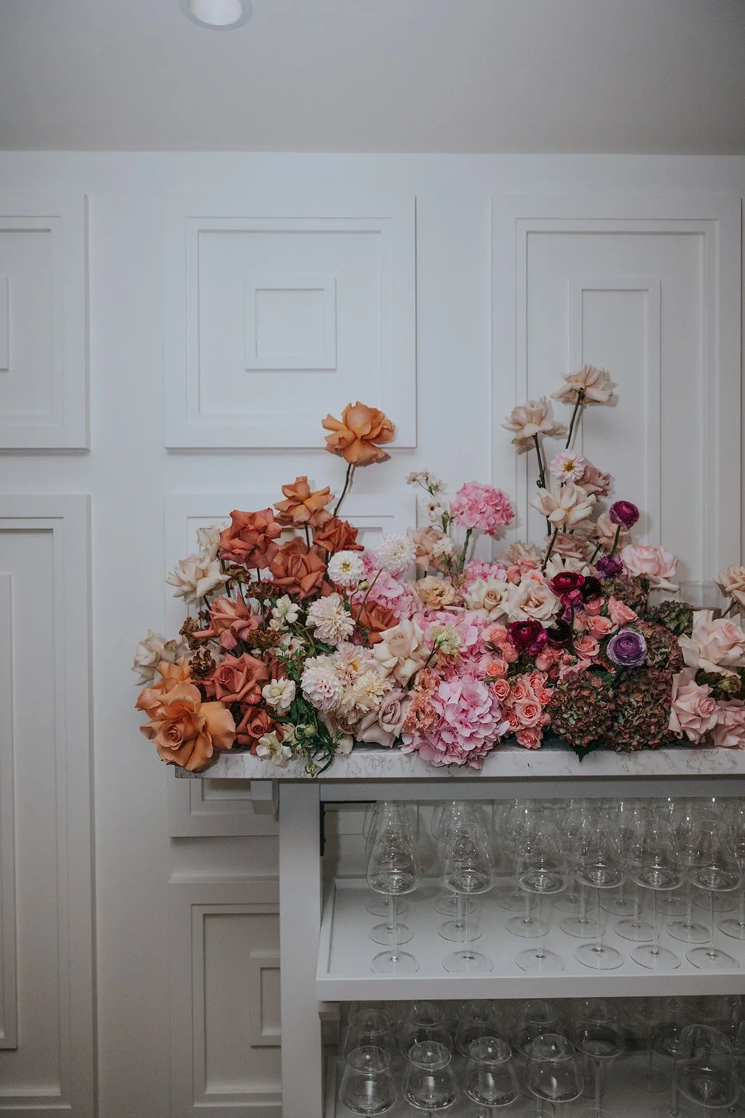 A floral arrangement with pink, peach, and purple flowers on a white table in a room with white paneled walls.