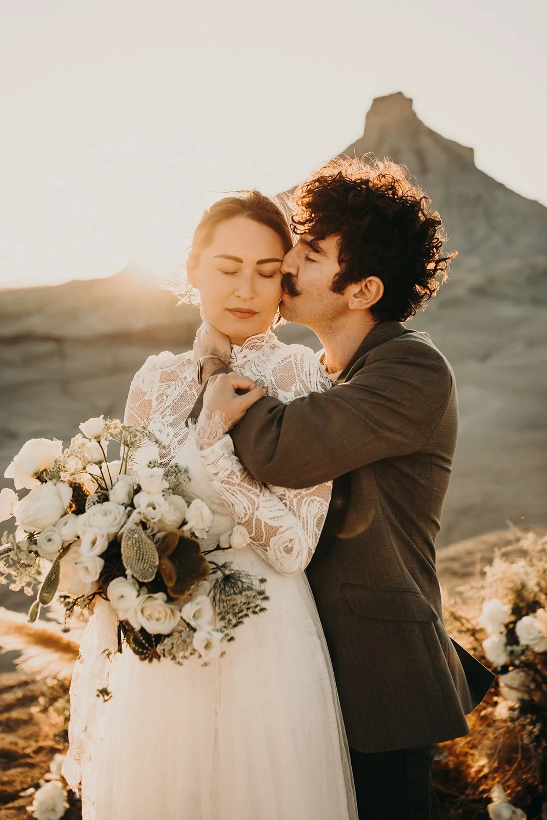 A couple on their wedding day embracing outdoors at sunset, with mountains in the background. The bride is holding a bouquet and has her eyes closed, while the groom kisses her cheek.