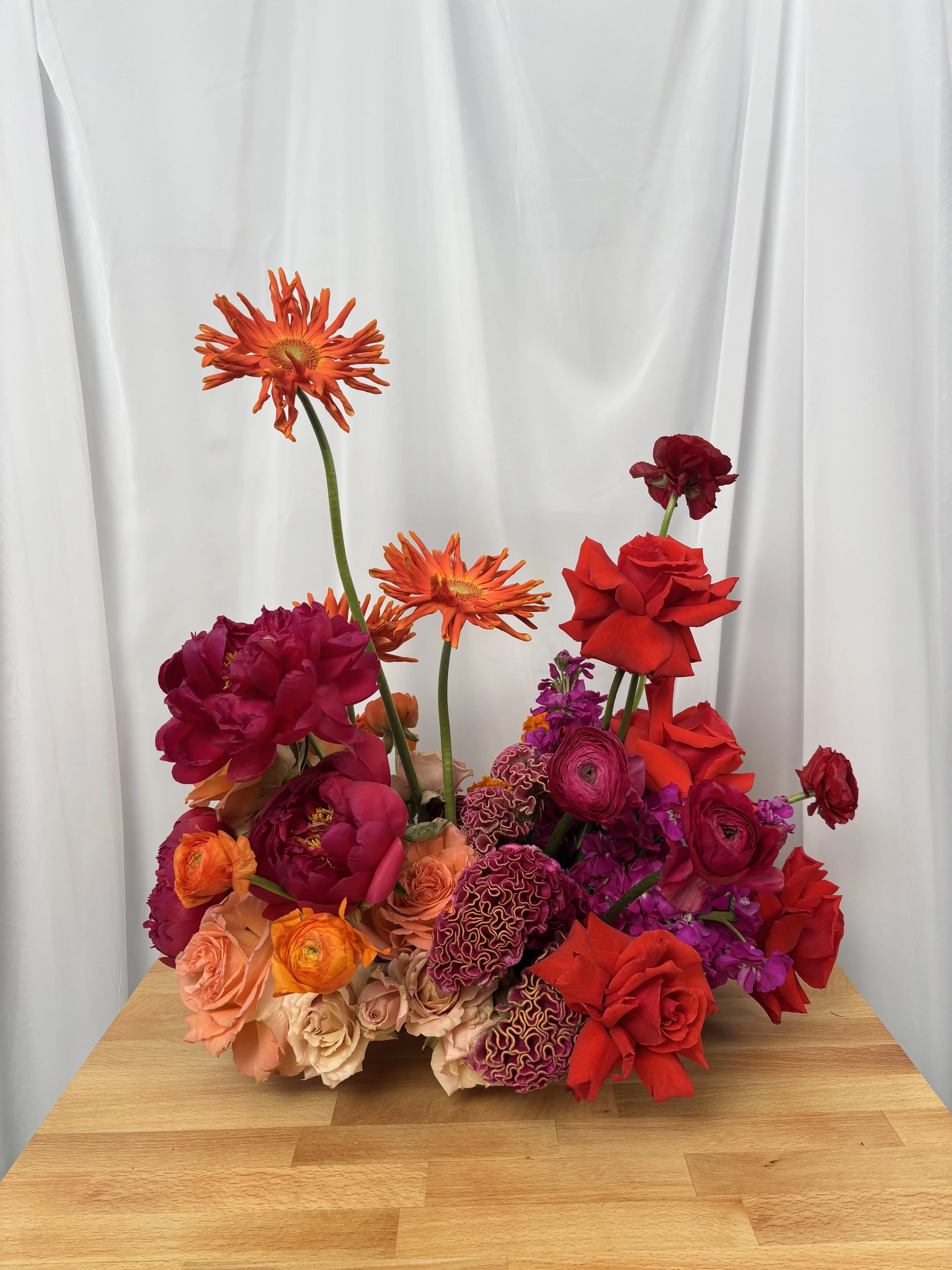 Colorful mixed flower arrangement with roses, dahlias, and other blooms on a wooden surface with white curtain backdrop.