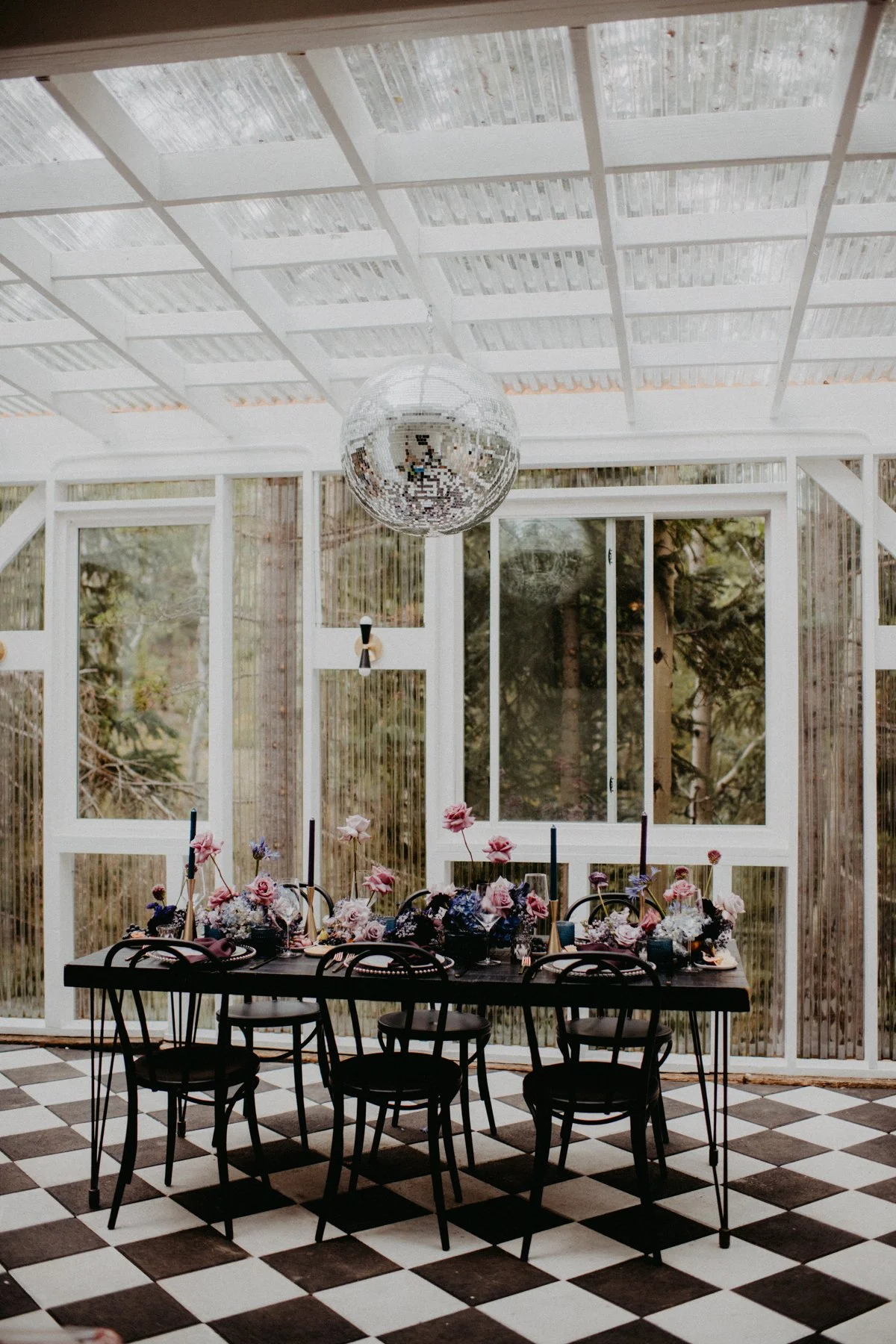 A dining table decorated with flowers, candles, and tableware in a glass-enclosed sunroom with large windows, a checkered black-and-white floor, and a disco ball hanging from the ceiling.