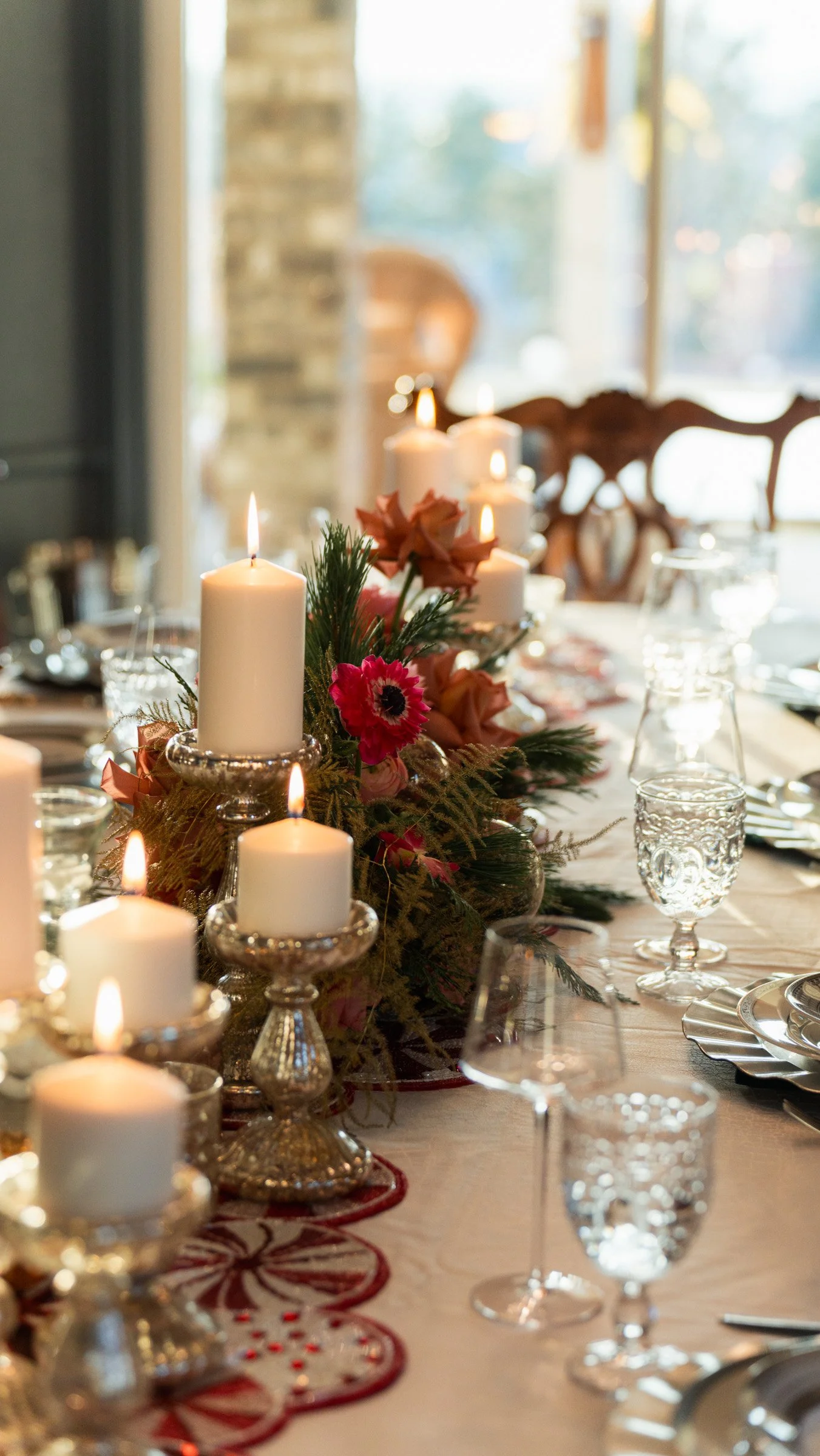 A decorated dining table with lit candles, floral arrangements, glassware, and plates, set near large windows with natural lighting.