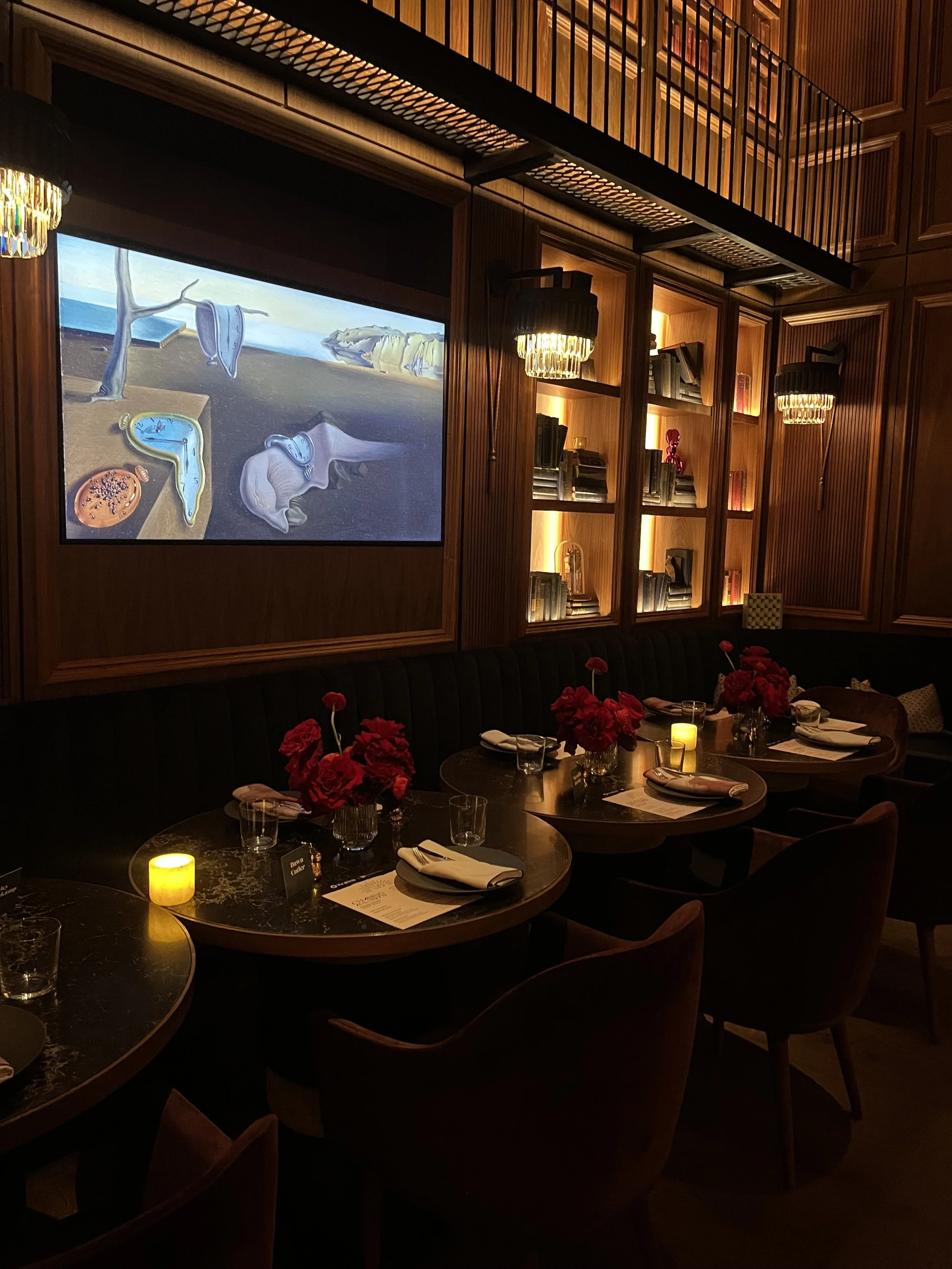 Elegant restaurant with dark wood paneling, round tables set with napkins, glasses, and flowers, dim lighting, a large screen displaying a surrealist image of melting clocks, and bookshelves with lighting.