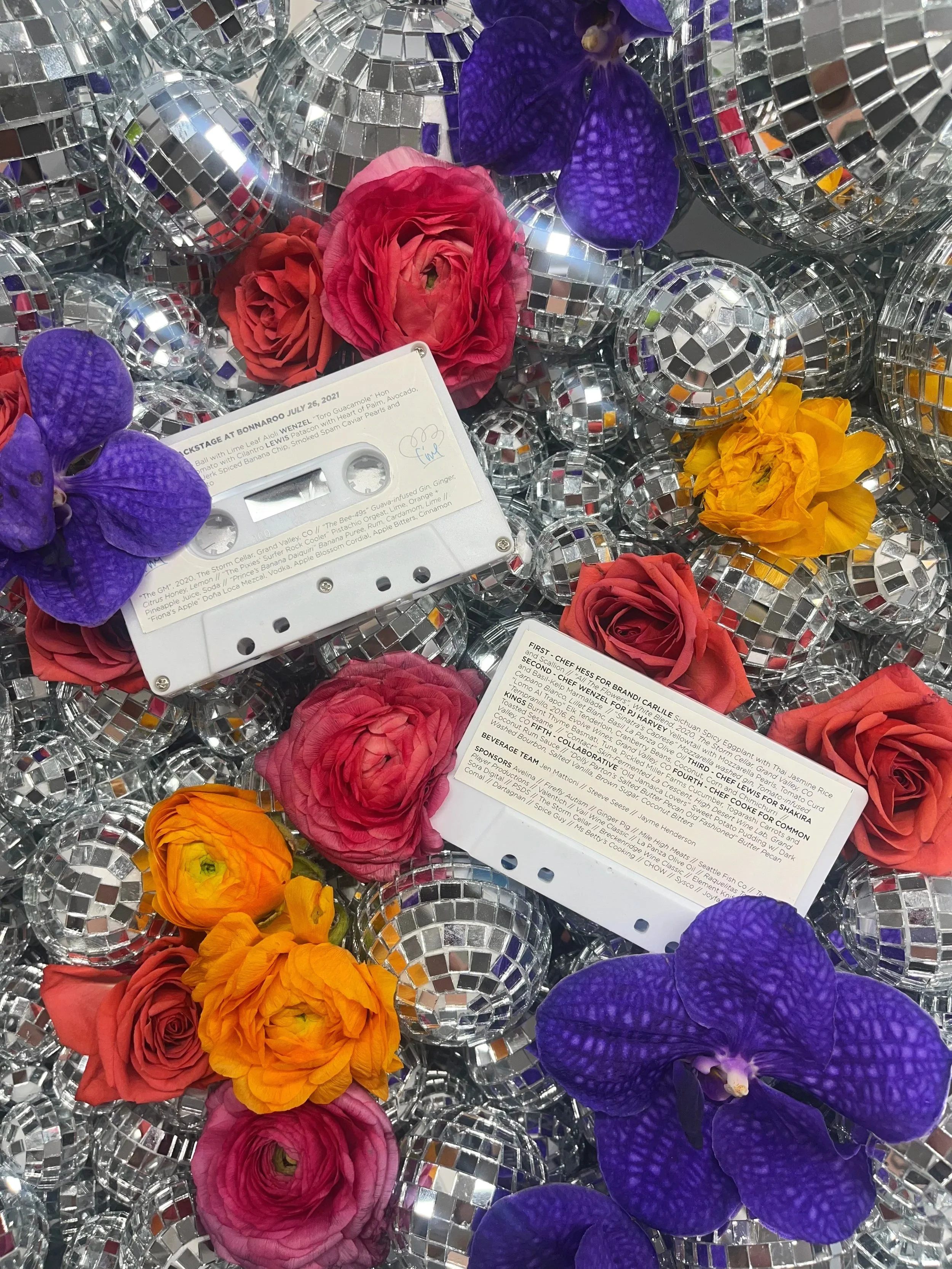 A collection of shiny disco balls intertwined with colorful flowers including purple orchids, pink roses, orange ranunculus, and red roses, with two cassette tapes placed among the items.