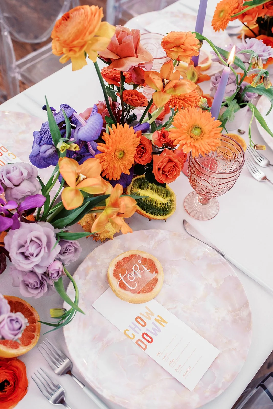 Table setting with a floral centerpiece, pink and purple glassware, cutlery, and a menu card on a pink marbled plate.