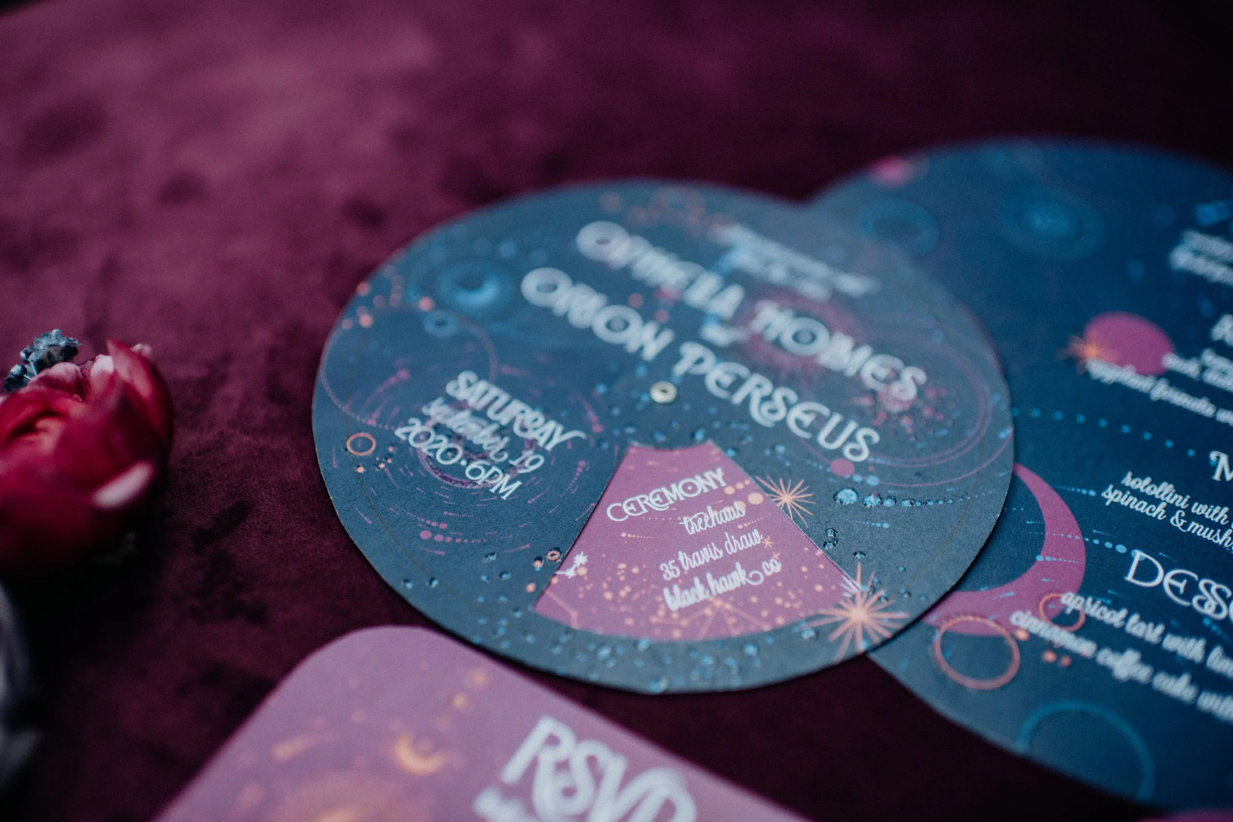 Wedding invitation cards with celestial and cosmic design, featuring text about the wedding date and ceremony details, on a dark purple surface with a small pink flower beside them.