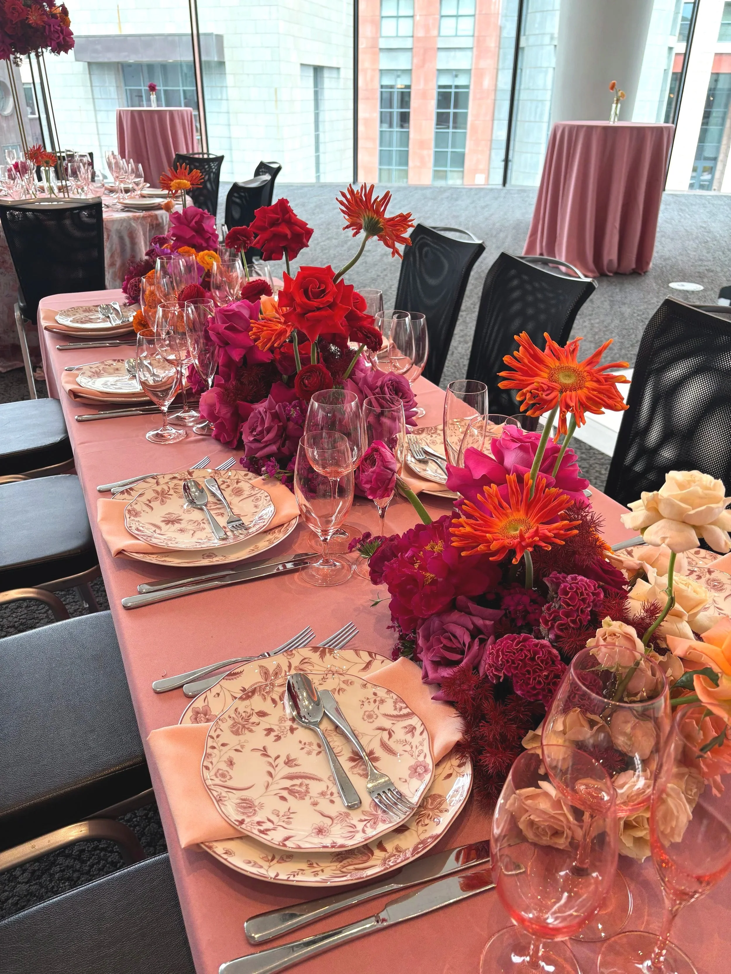 Elegant dining table set with pink tablecloth, floral centerpiece, fine china, glassware, and silverware, in a modern high-rise interior.