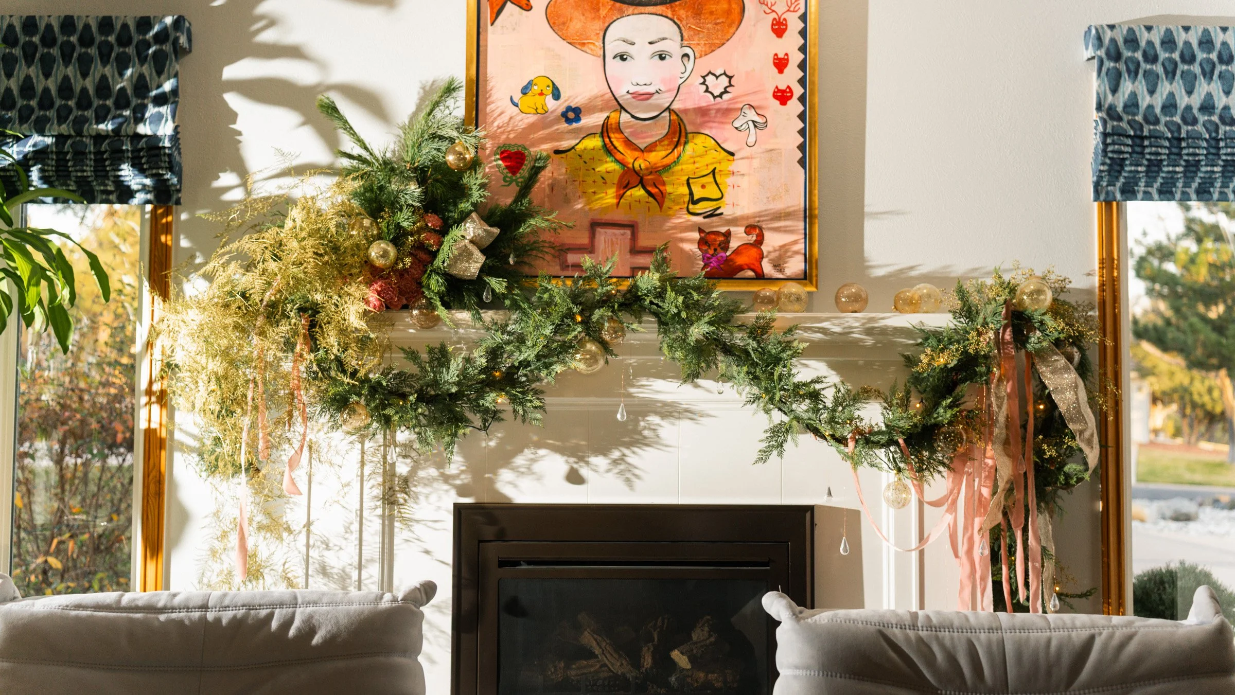 A decorated living room fireplace mantle with Christmas greenery, gold ornaments, and pink ribbons, set against a white wall with a colorful framed art piece above, and two large windows with blue patterned curtains flanking the scene.