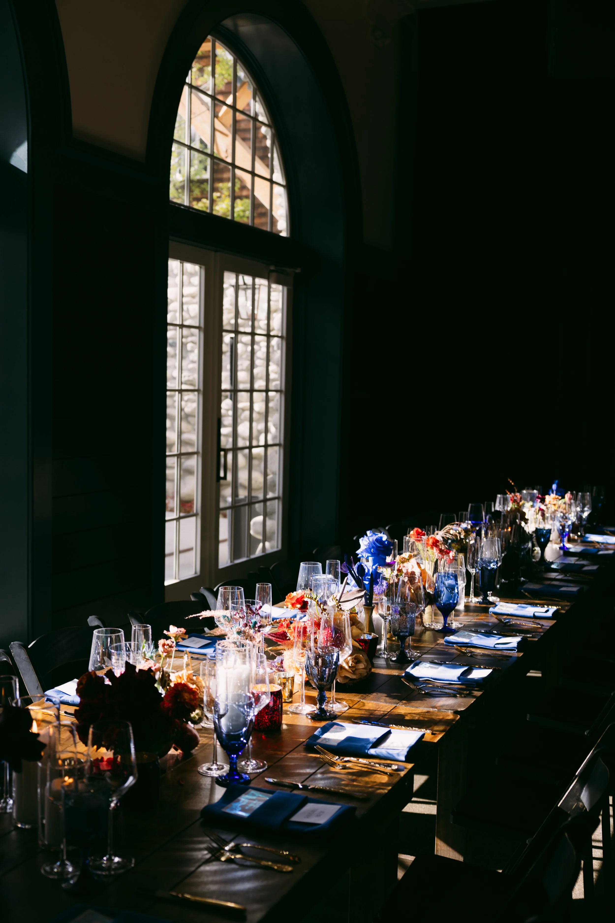 A long banquet table set with floral centerpieces, blue and clear glassware, and tableware, illuminated by sunlight through large arched windows in a dark room.