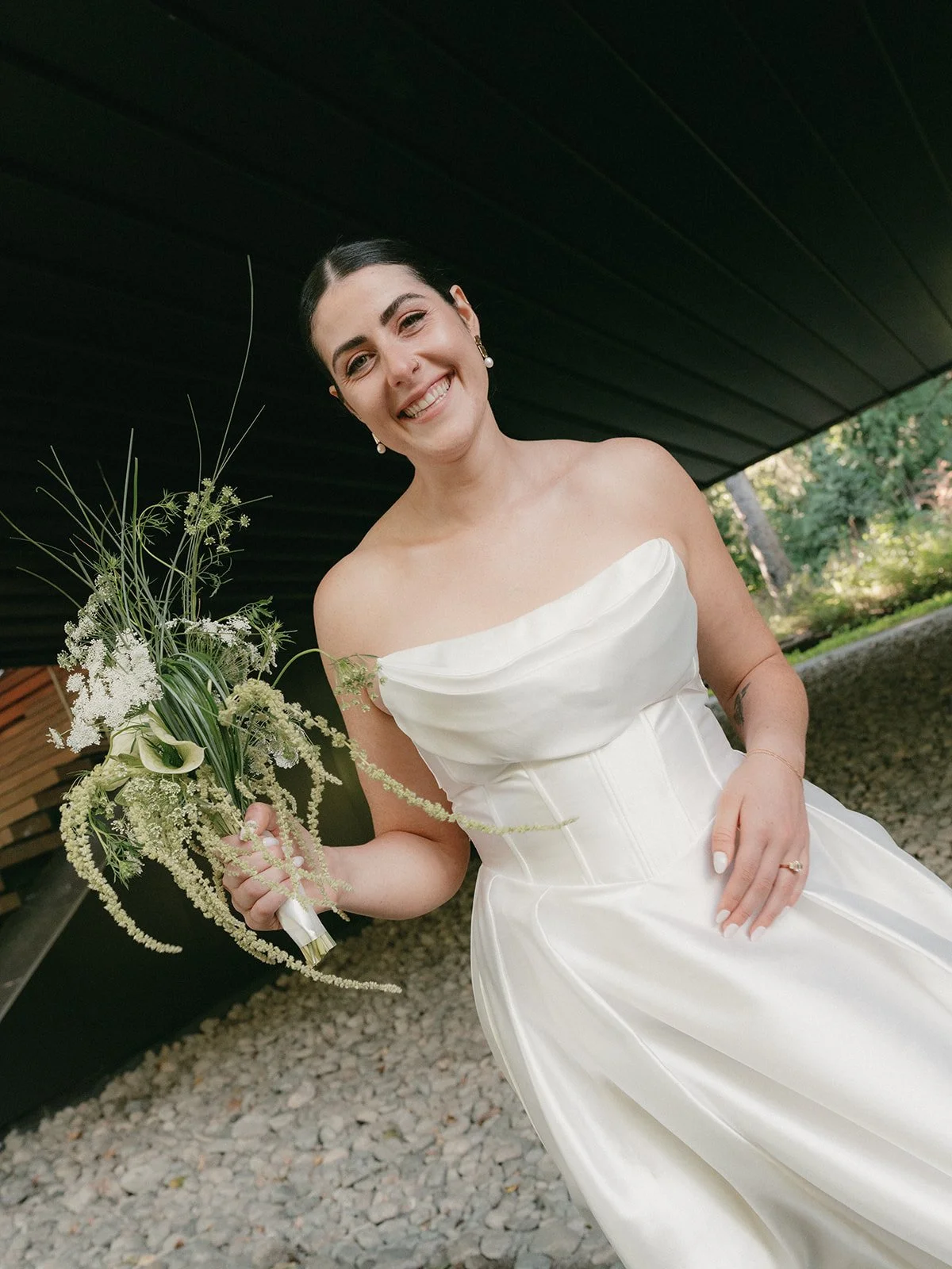 A smiling woman in a white wedding dress holding a bouquet of flowers outside near a dark building and trees.