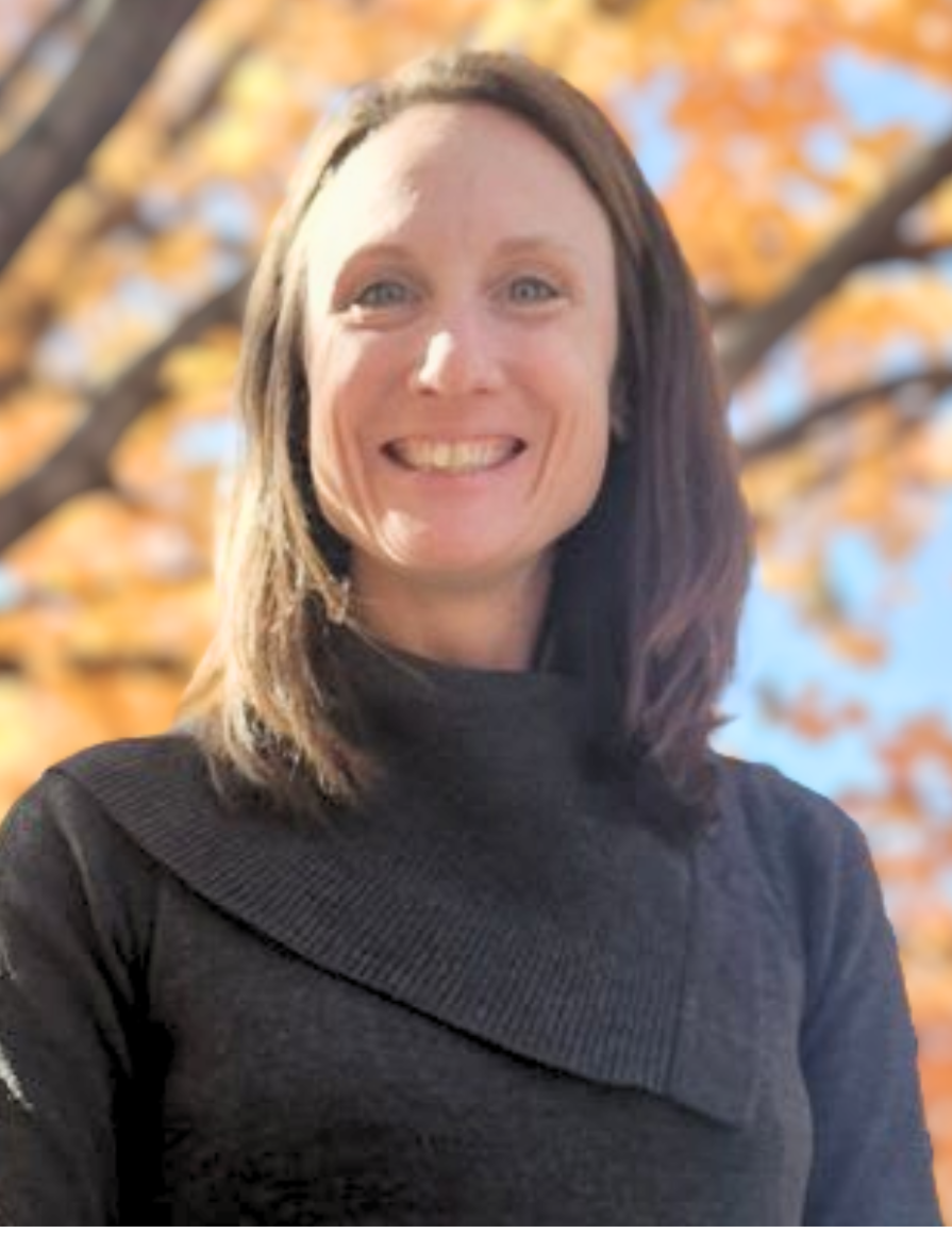 A woman with brown hair and blue eyes smiling outdoors with autumn-colored leaves in the background.