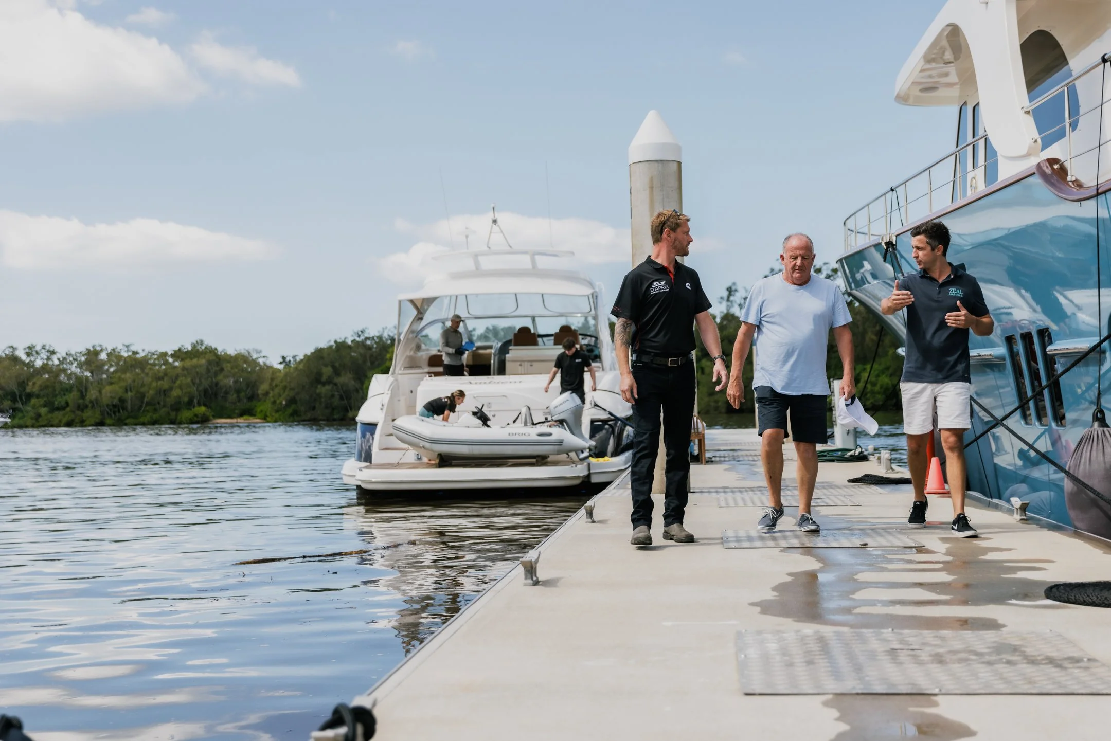 People walking on a boat dock near boats, including a yacht and a smaller boat, on a calm body of water with trees in the background and a partly cloudy sky.