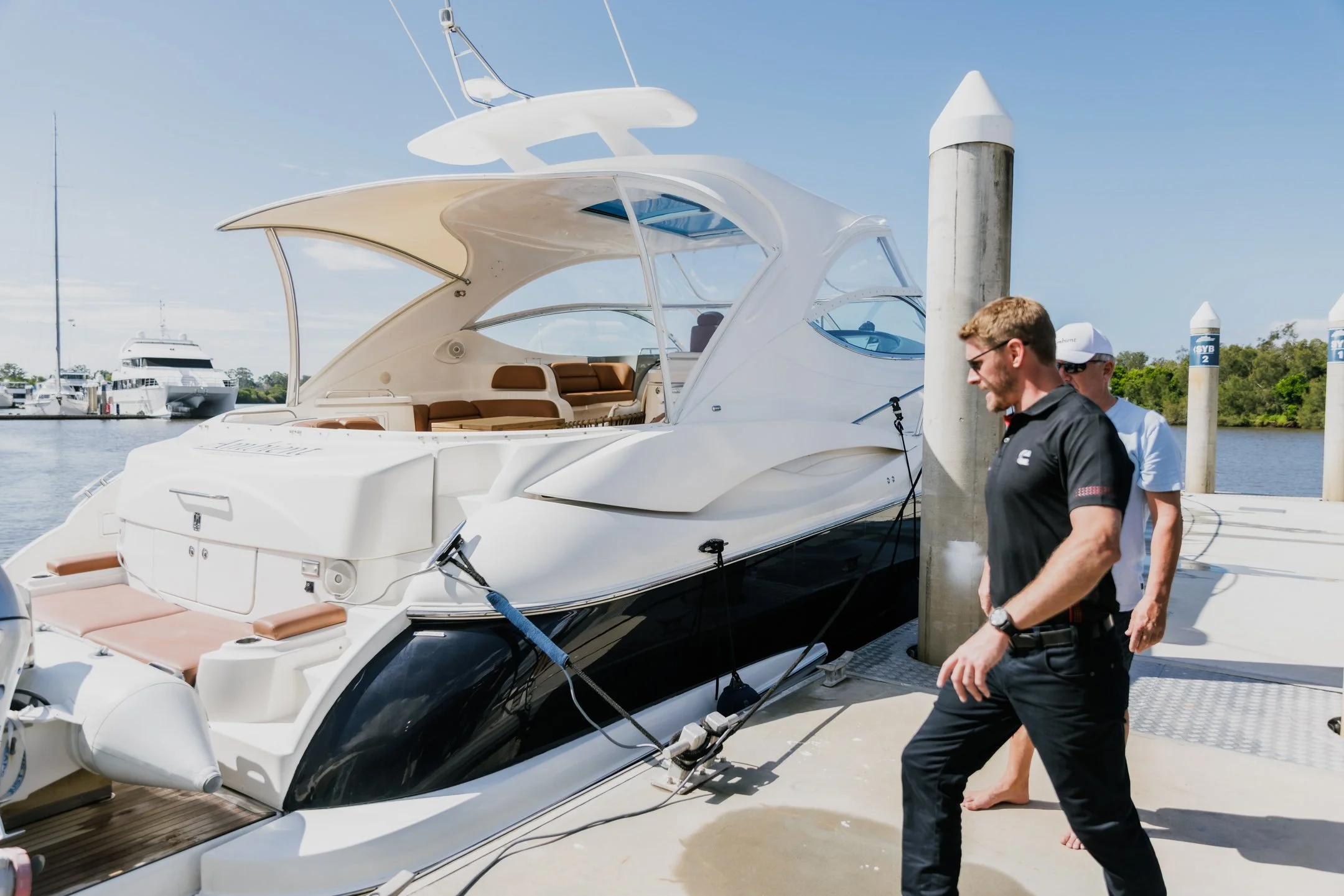 A white yacht docked at a marina with two men walking nearby on the dock. One man is wearing sunglasses and a black shirt, and the other is wearing a white shirt and a white cap. The background includes water, other boats, and a clear sky.