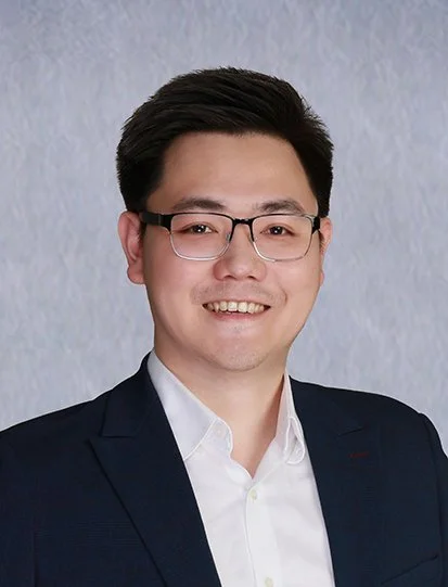 A smiling man wearing glasses, a white shirt, and a dark suit jacket against a plain light blue background.
