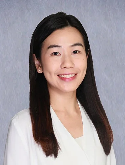 A young woman with long black hair, smiling, wearing a dark blue top, standing against a plain light-colored background.