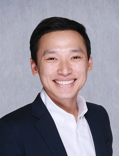 A young man smiling, wearing a white button-up shirt, standing against a plain light-colored wall.