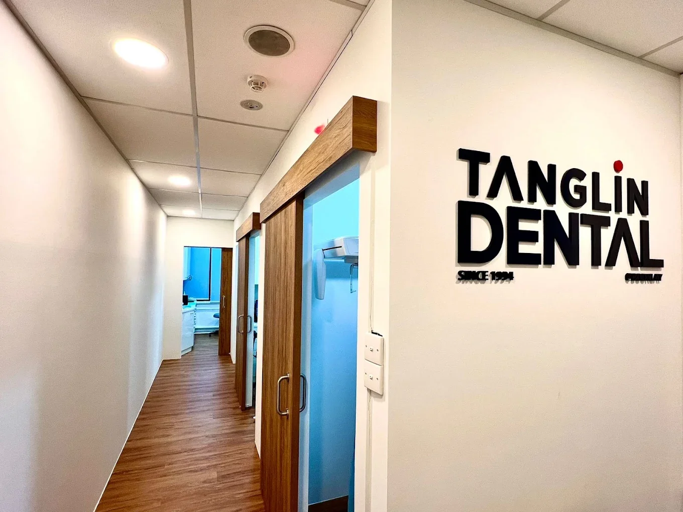 Interior view of a dental office corridor with wooden sliding doors, a white wall with a sign that reads "TANGLIN DENTAL," and a dental X-ray machine visible through the open door.