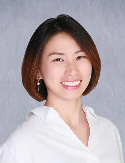 A woman with shoulder-length brown hair smiling, wearing a white top with lace sleeves, standing indoors.