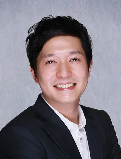 Professional portrait of a young Asian man in a dark suit and white shirt, smiling, against a neutral light gray background.